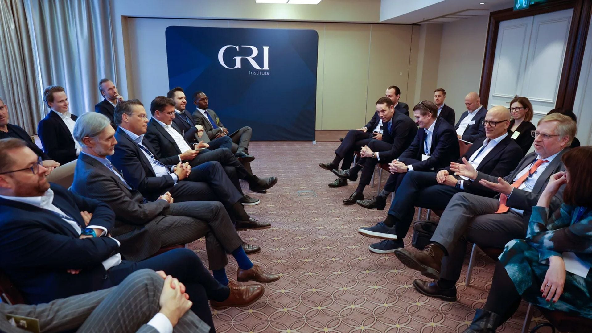 A professional group of people in business attire is seated in two facing rows in a conference room, engaged in a discussion, likely concerning real estate investments or strategy in Germany. A large banner with the "GRI Institute" logo is visible at the back of the room.