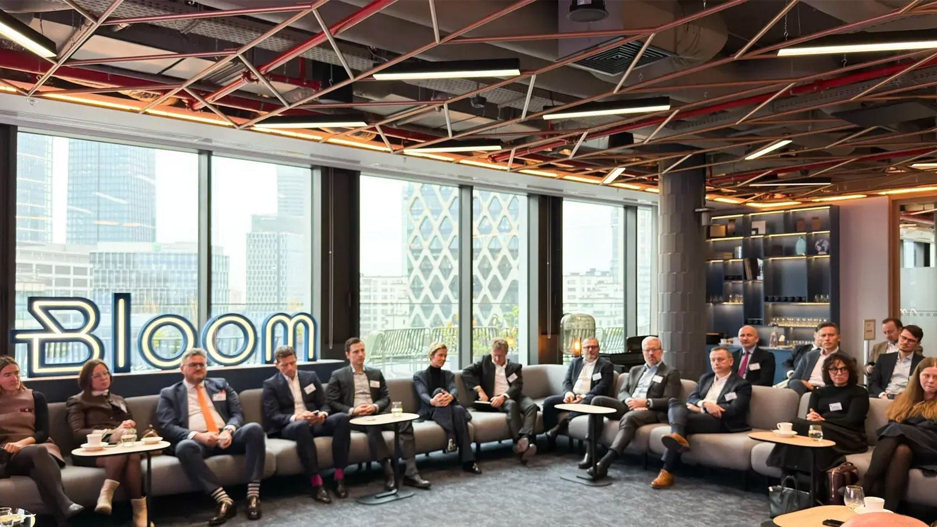 An image showing a group of real estate leaders and economists seated in a semi-circle during a GRI Institute roundtable discussion in a modern office. The room features floor-to-ceiling windows overlooking a city skyline, a "Bloom" neon sign, and an industrial-style ceiling.