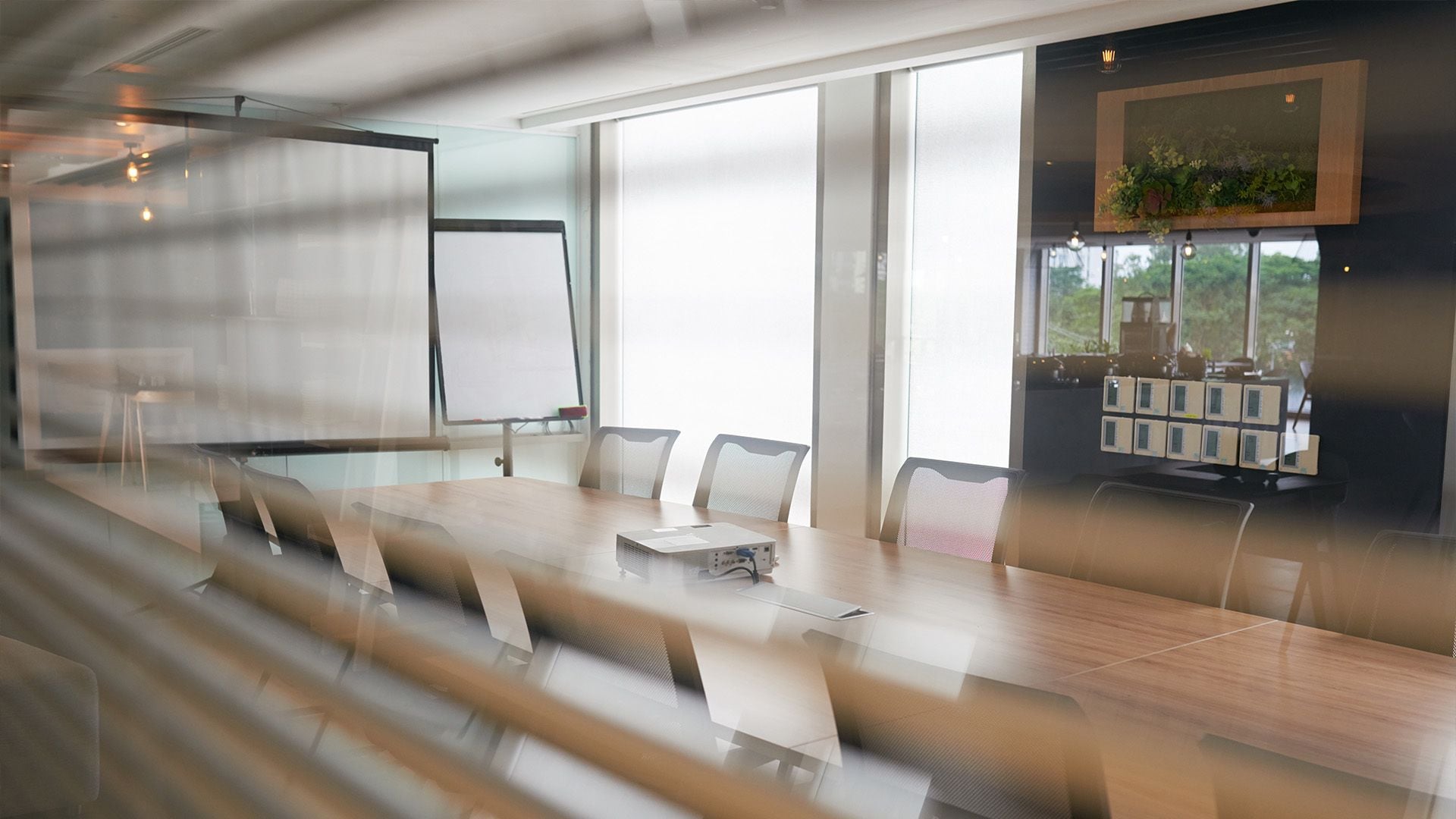 An interior view of a premium real estate development showcasing a collaborative office space with a long meeting table, projector, whiteboard, and biophilic wall art behind glass window blinds.