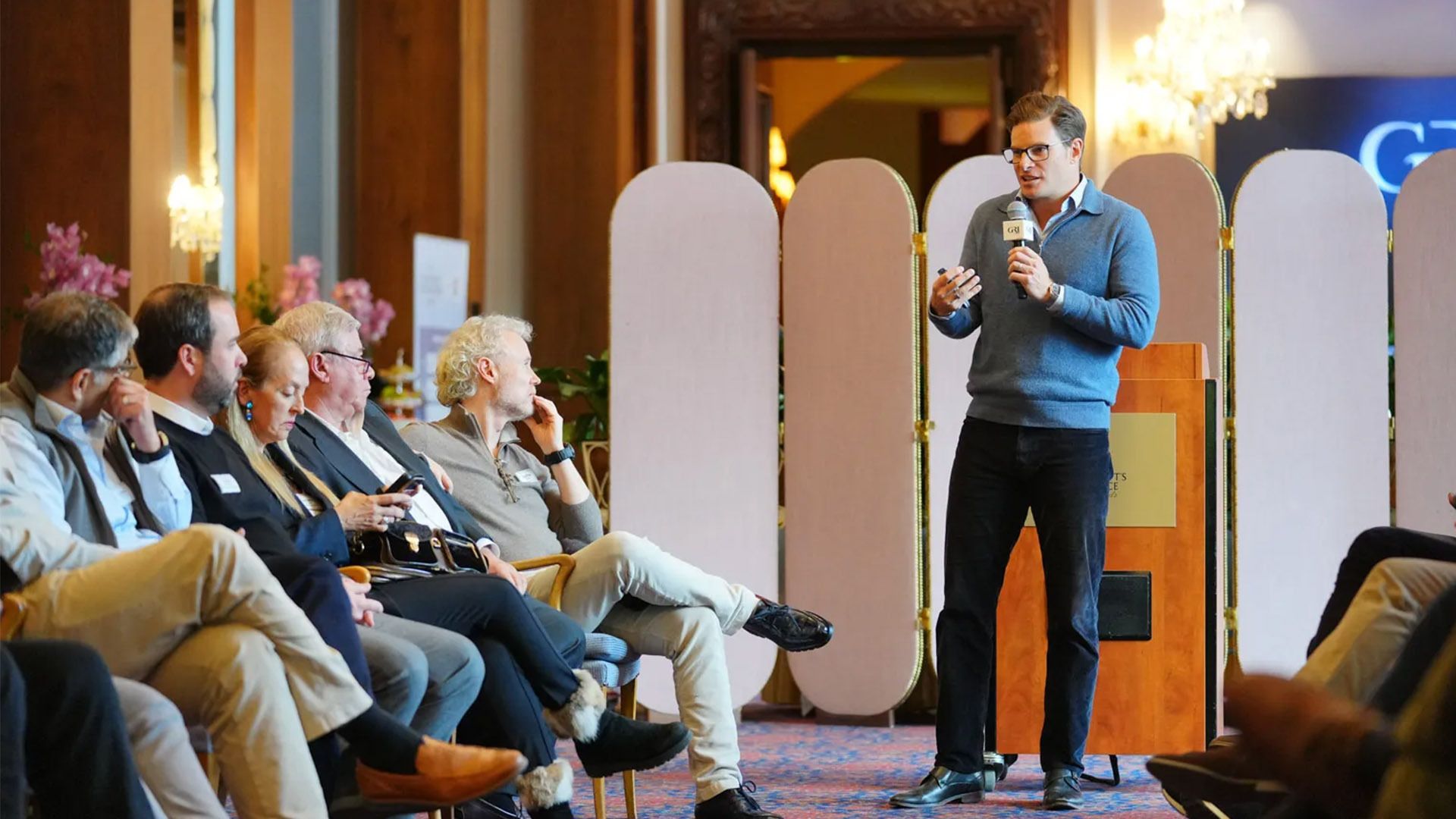 David Stubbs addresses a group of seated attendees at the GRI Chairmen's Retreat Europe 2026. Stubbs, wearing a blue quarter-zip sweater and glasses, stands at a wooden podium holding a microphone and a clicker. The audience is seated in a row of chairs against a backdrop of folding privacy screens in an elegant indoor venue.