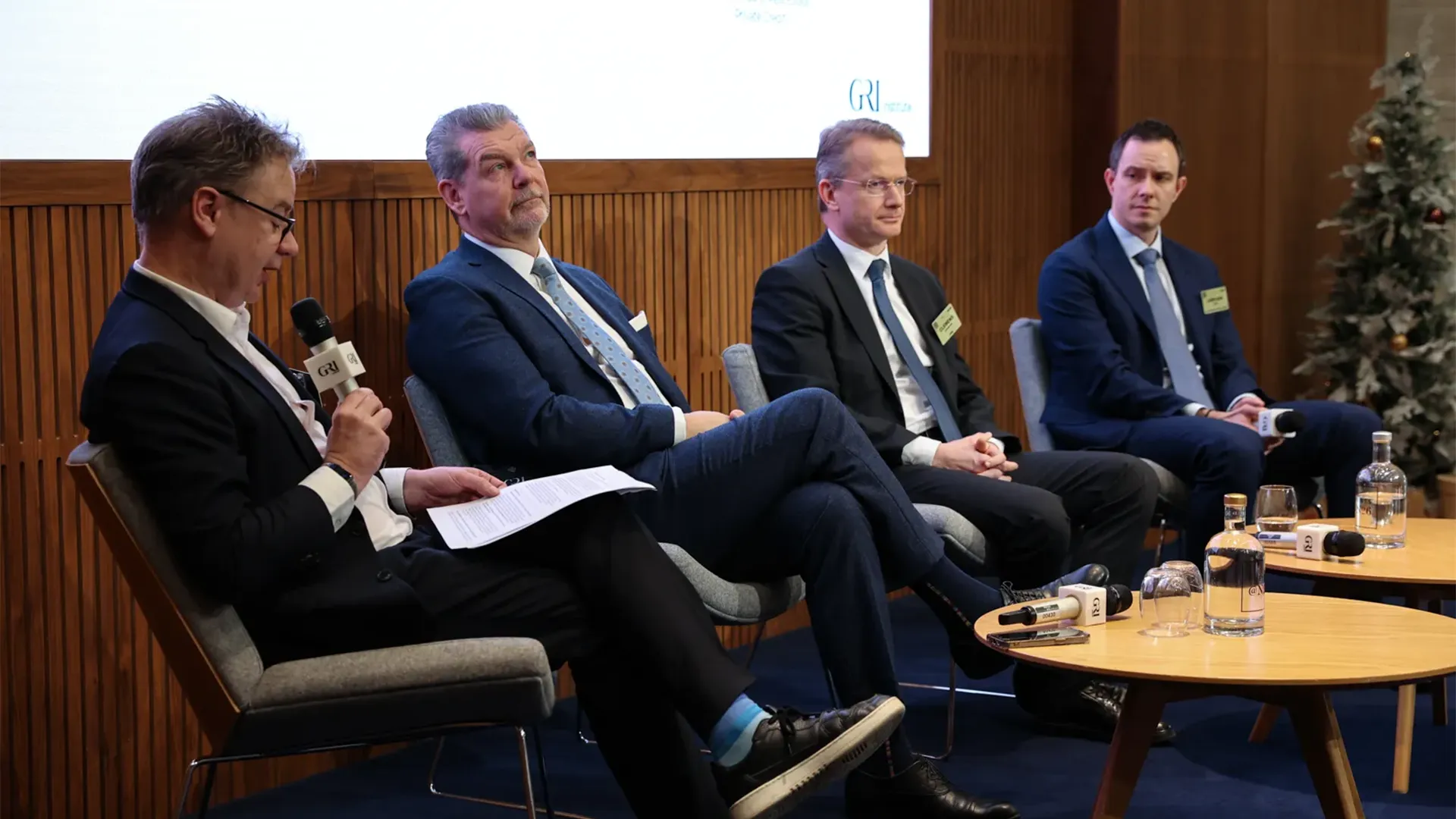A panel discussion on a stage with four men in business suits. The man on the far left, wearing glasses, is speaking into a microphone and holding papers. The other three men are seated and listening, with two of them looking towards the speaker. The setting appears to be a conference or event focused on the real estate sector, with wood-paneled walls and a small decorated Christmas tree visible in the background on the right.