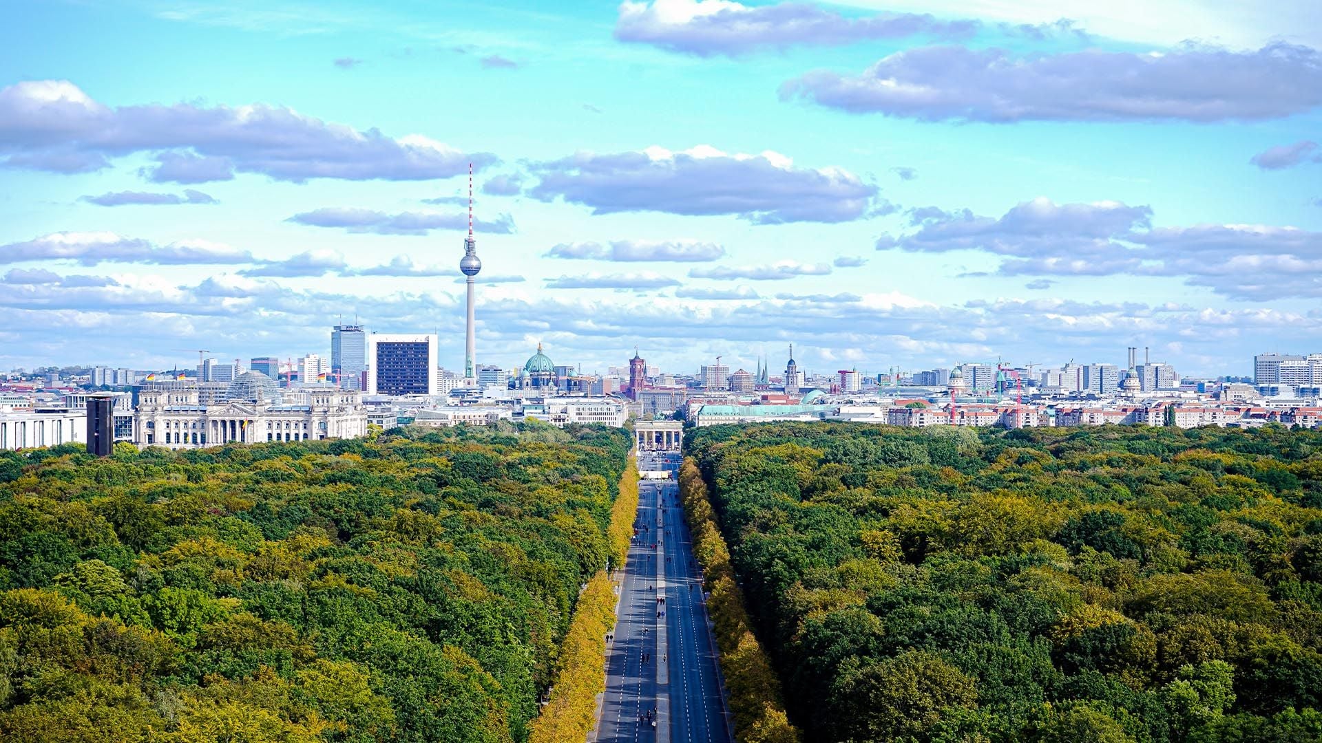 featuring the Tiergarten park in the foreground, with the Straße des 17. Juni cutting through the lush green trees toward the Brandenburg Gate and the Berlin TV Tower (Fernsehturm) under a bright, cloudy blue sky.