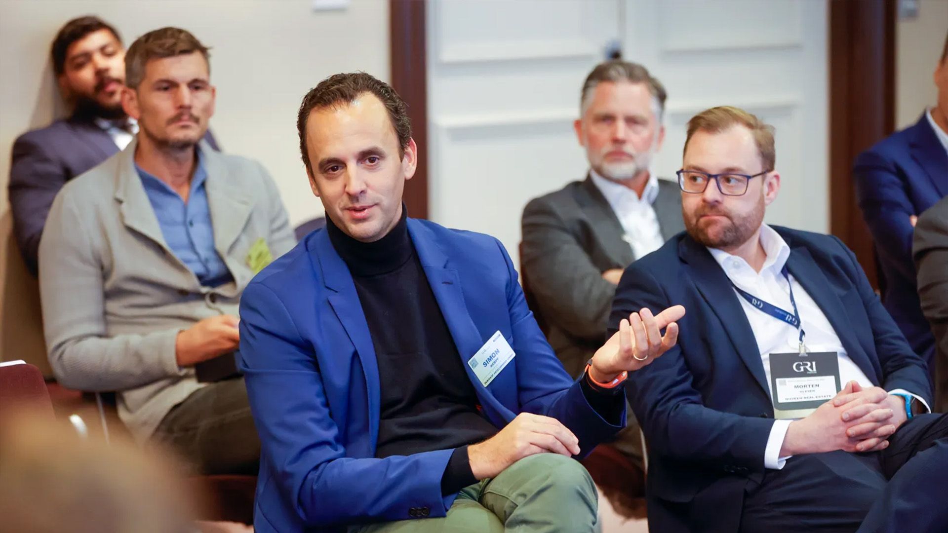 A man wearing a bright blue jacket and a black turtleneck is sitting and actively speaking during a conference or panel discussion, likely about the real estate market in Germany. He is identified by his name badge as "MARCO." Several other male attendees are seated around him, listening.