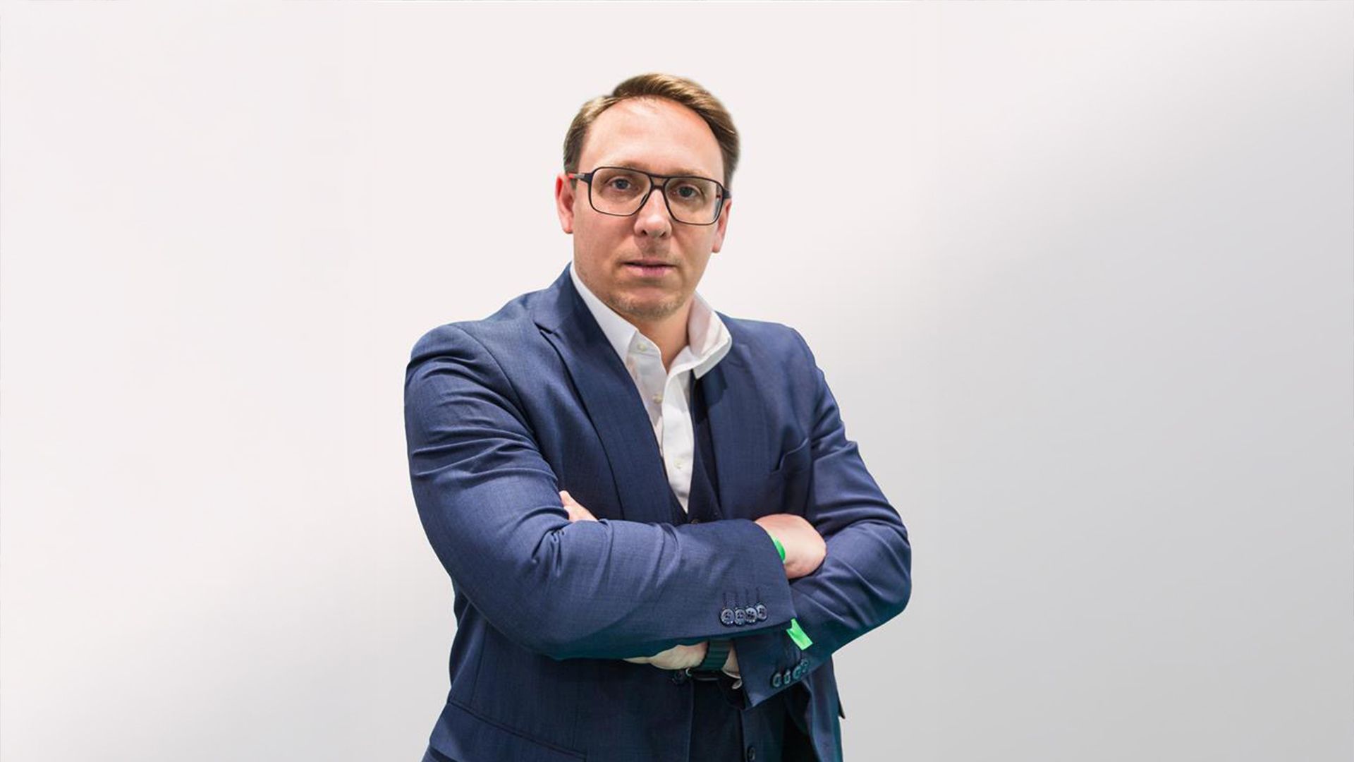 A professional portrait of Federico Valentini from Nhood, wearing a blue tailored suit, white shirt, and glasses, standing with arms crossed against a neutral white background.