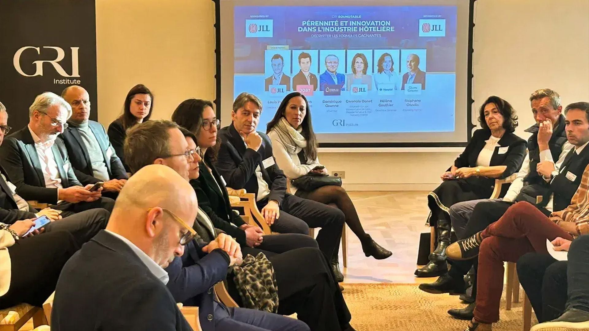 An image showing a panel discussion on real estate and hospitality at a GRI Institute event, featuring attendees and speakers seated in a room, with a screen in the background.