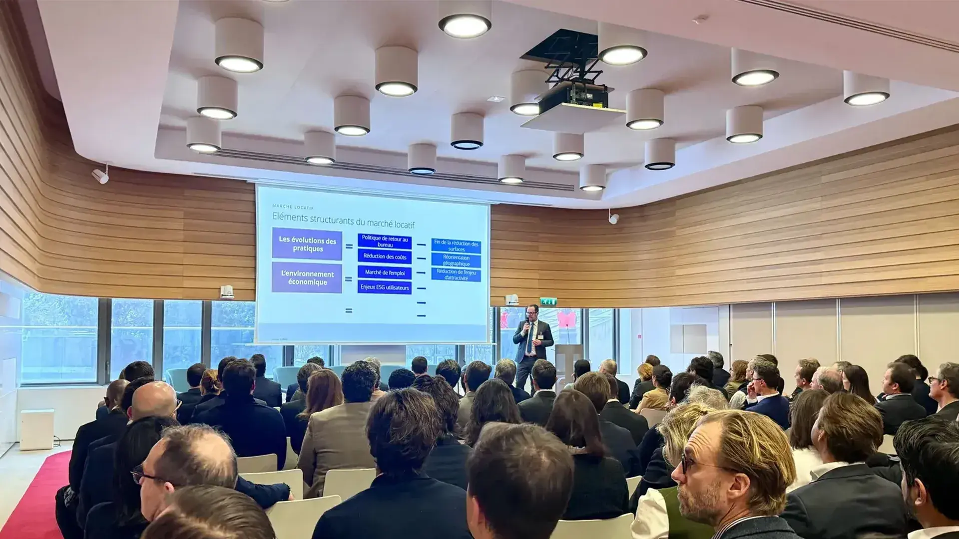 A wide shot of a French real estate conference. A presenter stands at the front of a wood-paneled room next to a projection screen titled "Marché Locatif" (Rental Market). The audience, seen from behind, listens as the presentation outlines structural elements of the market, including "L'environnement économique" and "Les évolutions des pratiques."
