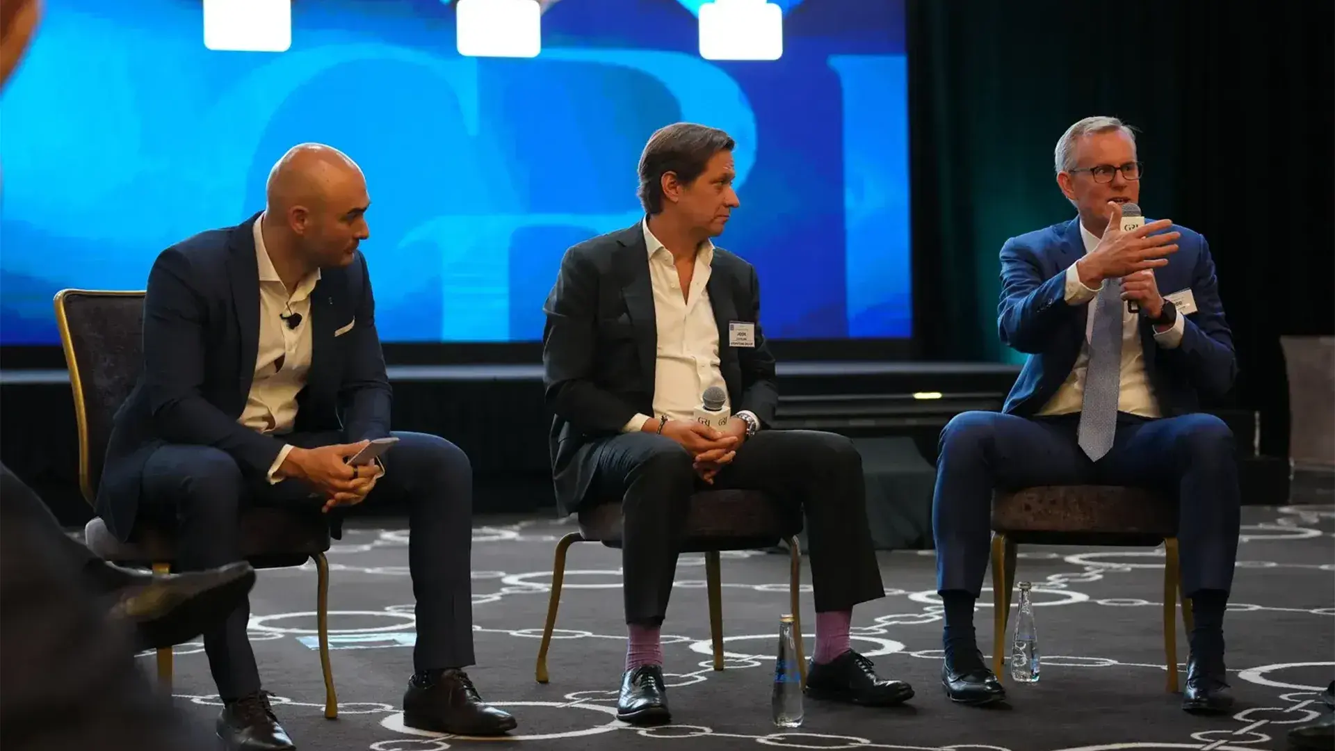 A panel of three global real estate leaders dressed in business suits sits on stage, engaged in discussion. The man on the right is speaking into a microphone.