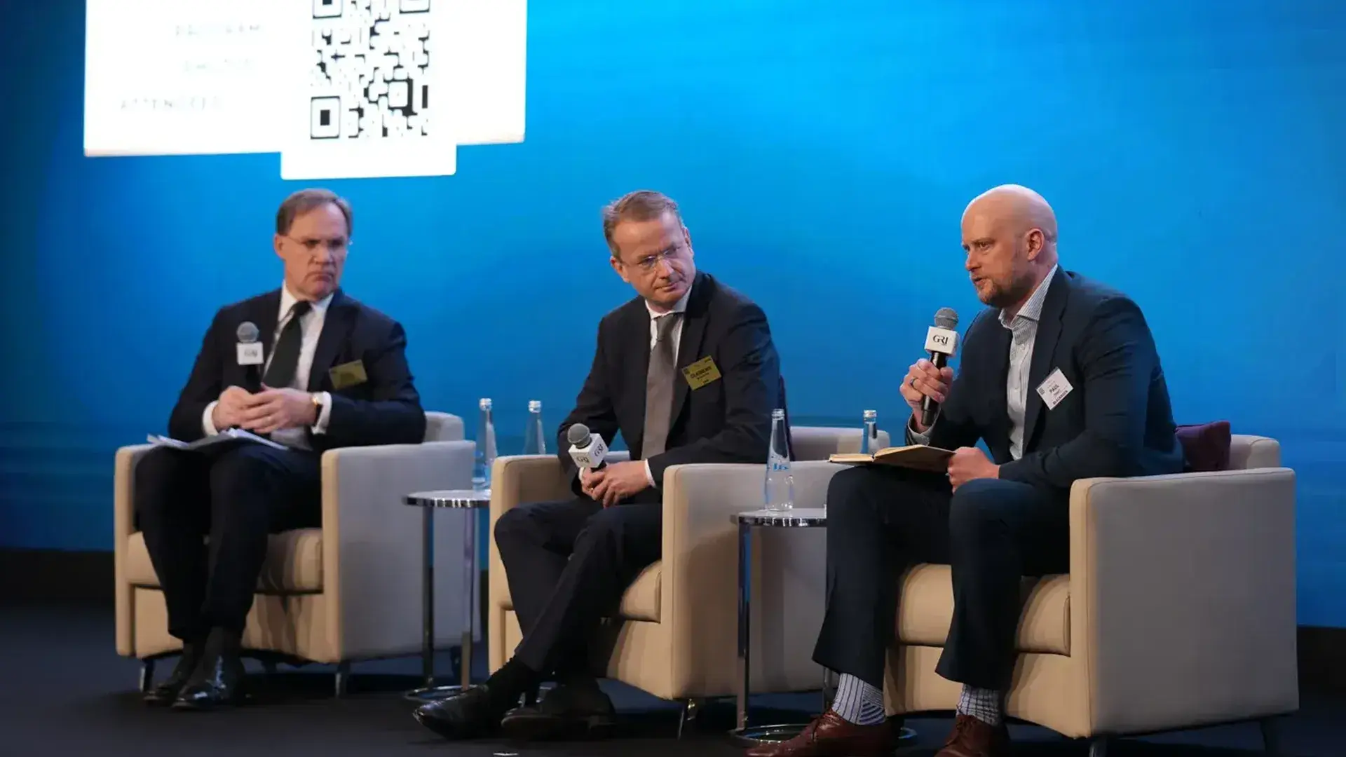 Three men in business suits participate in a panel discussion at a global real estate summit. The man on the right holds a microphone and a notebook, speaking to the seated audience.