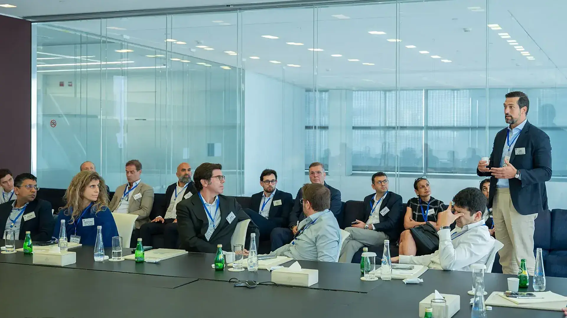 A professional man in a beige and blue suit is presenting and speaking to a group of approximately a dozen executives and investors seated around a large table in a modern conference room. The meeting is likely taking place at ADGM (Abu Dhabi Global Market) and focusing on investment or real estate development.