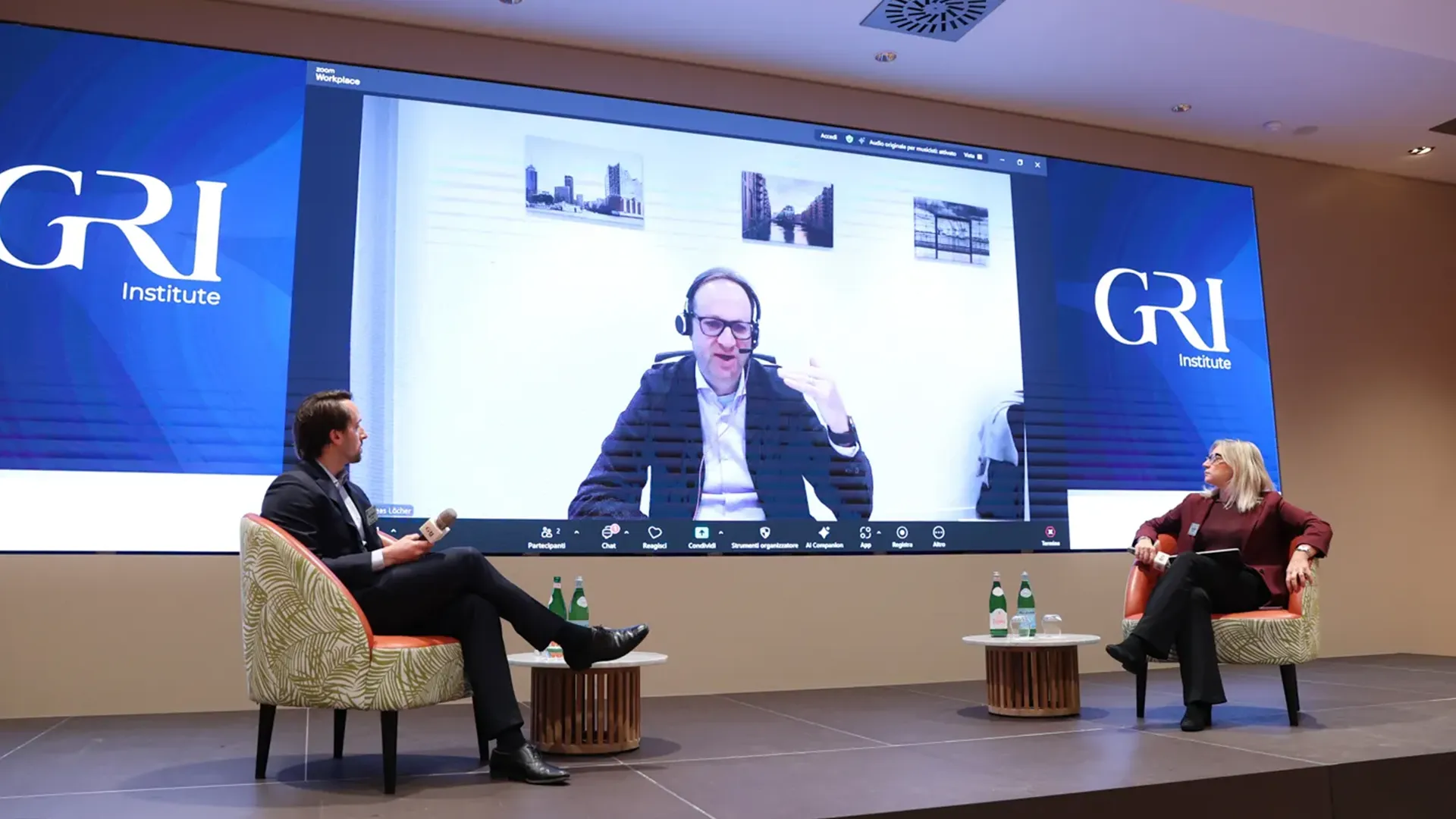Two speakers on stage, one man and one woman, participating in a panel discussion with a third speaker visible remotely on a large screen displaying the GRI Institute logo, addressing trends in the hospitality real estate sector.