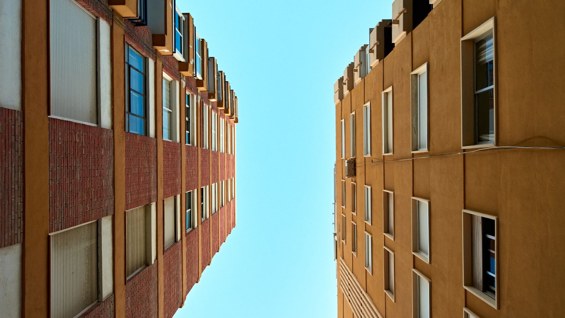 A low-angle, "worm's-eye view" looking directly up between two multi-story residential real estate buildings toward a clear, bright blue sky. On the left, a tall building features a facade of warm red brick and tan panels with vertical windows. On the right, a parallel building is finished in a solid ochre-yellow textured plaster with rows of white-trimmed windows. The two structures create a strong sense of symmetry and urban density, framing the sky in a narrow vertical corridor.