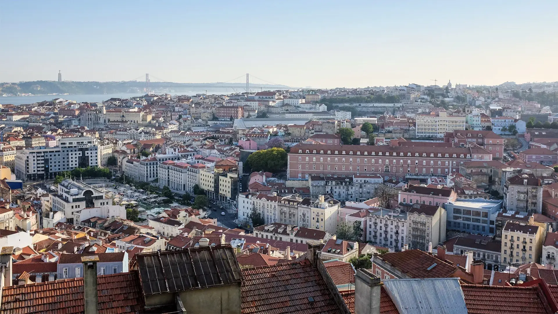 An elevated, wide-angle view of the Lisbon city skyline, showcasing a dense mix of historic architecture and modern real estate. The urban landscape features a variety of commercial offices and residential buildings with traditional terracotta roofs, leading toward the 25 de Abril Bridge and the Tagus River in the distance under a clear, bright sky.