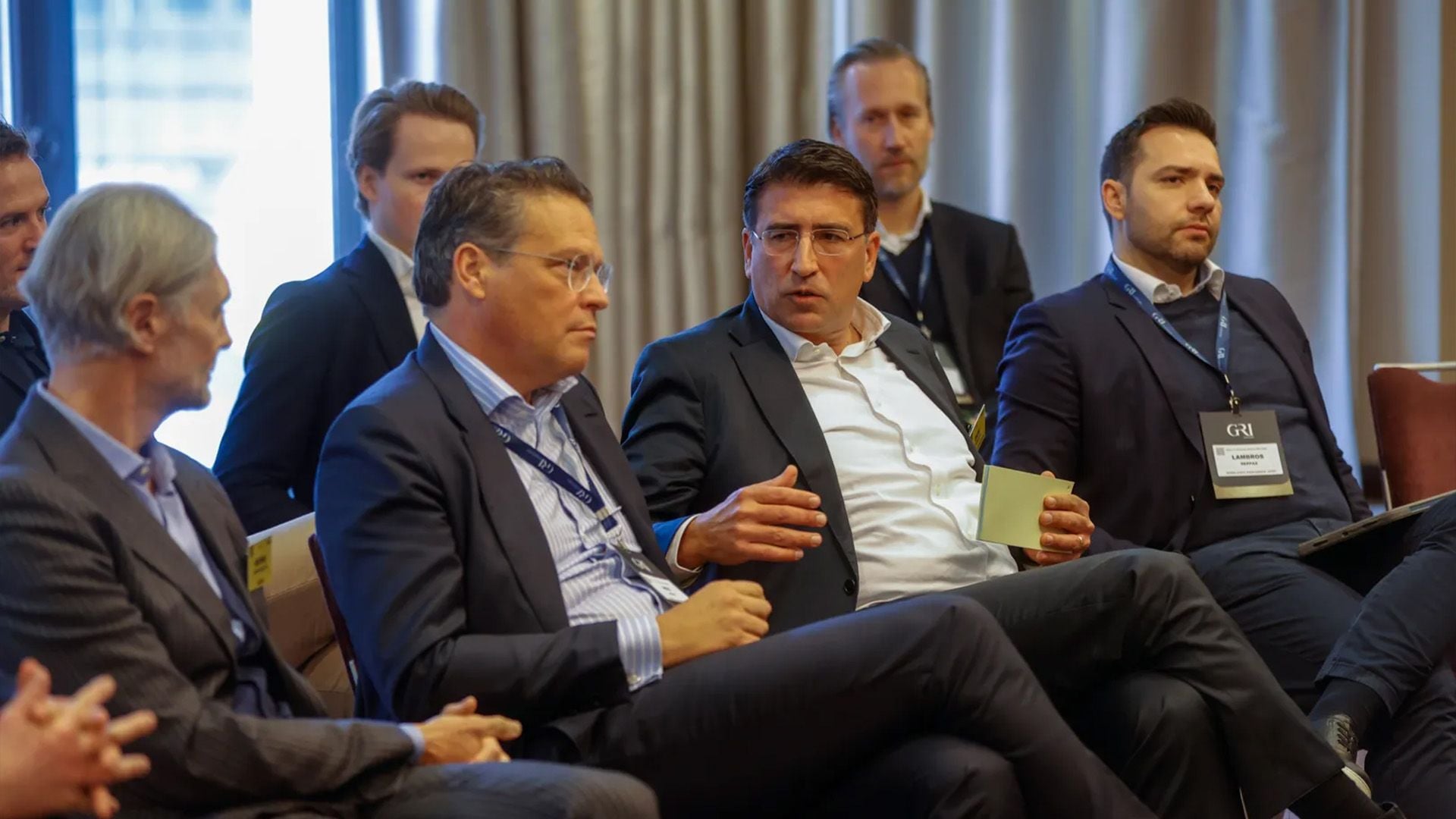 A close-up view of several male business professionals in suits attending a seated discussion, potentially a meeting or conference focused on real estate investment trends and opportunities in Germany. One man in the center is actively speaking while holding a small piece of paper.