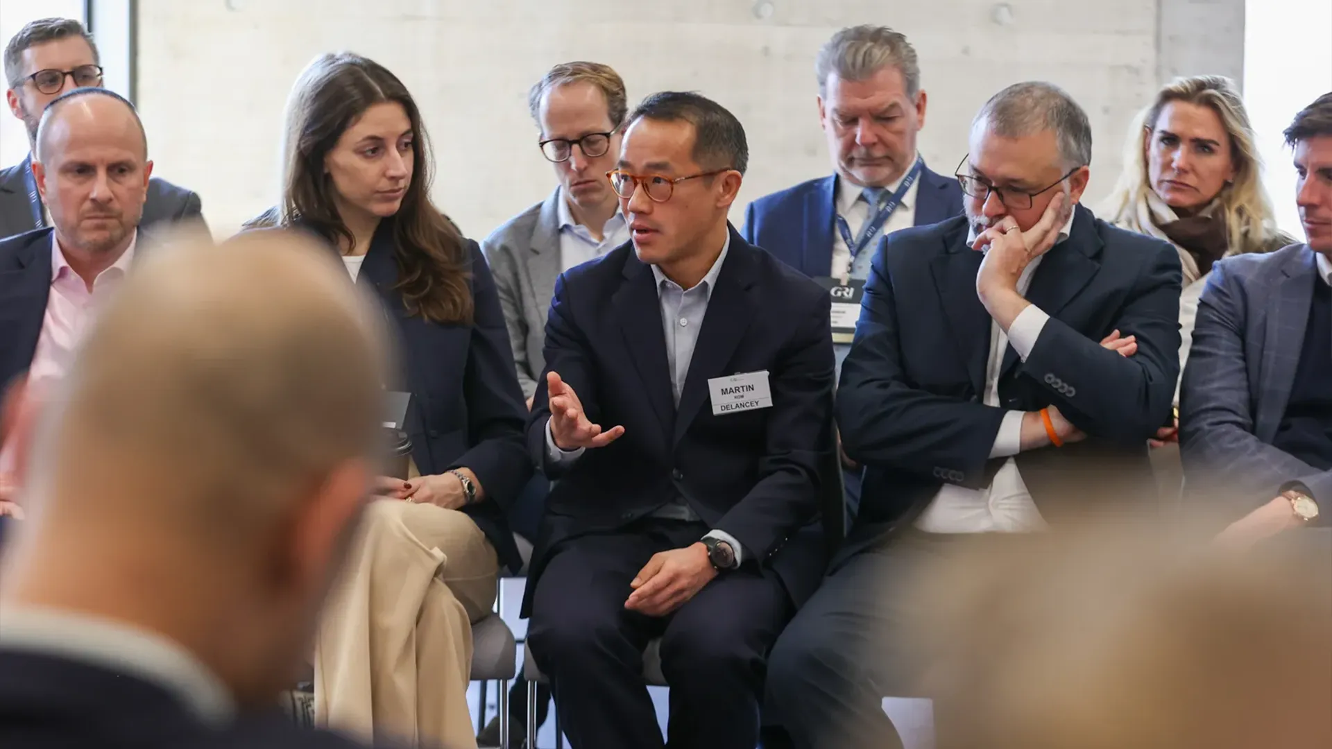 A candid photo of a group of business professionals sitting in a circle during a discussion or presentation. The focus is on a man in the centre, named MARTIN DELANEY according to his nametag, who is speaking animatedly with an open hand gesture. He is surrounded by several other attendees, both men and women, who are listening intently. The setting is modern, with a concrete wall visible in the background, suggesting a corporate or conference environment, perhaps about real estate.