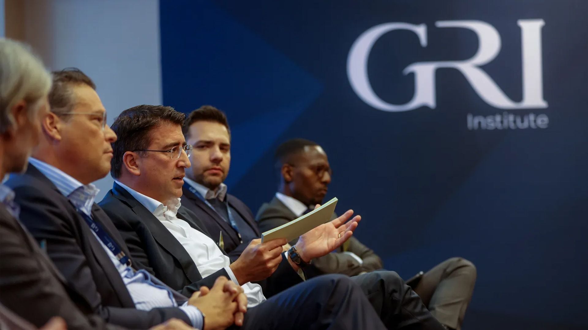 A panel discussion in front of a GRI Institute backdrop, featuring four men in suits. The man in the center is speaking and gesturing with a document in his hand. The image captures a professional discussion likely related to real estate and possibly focusing on Germany, given the context of the GRI Institute's global focus on sustainability reporting.