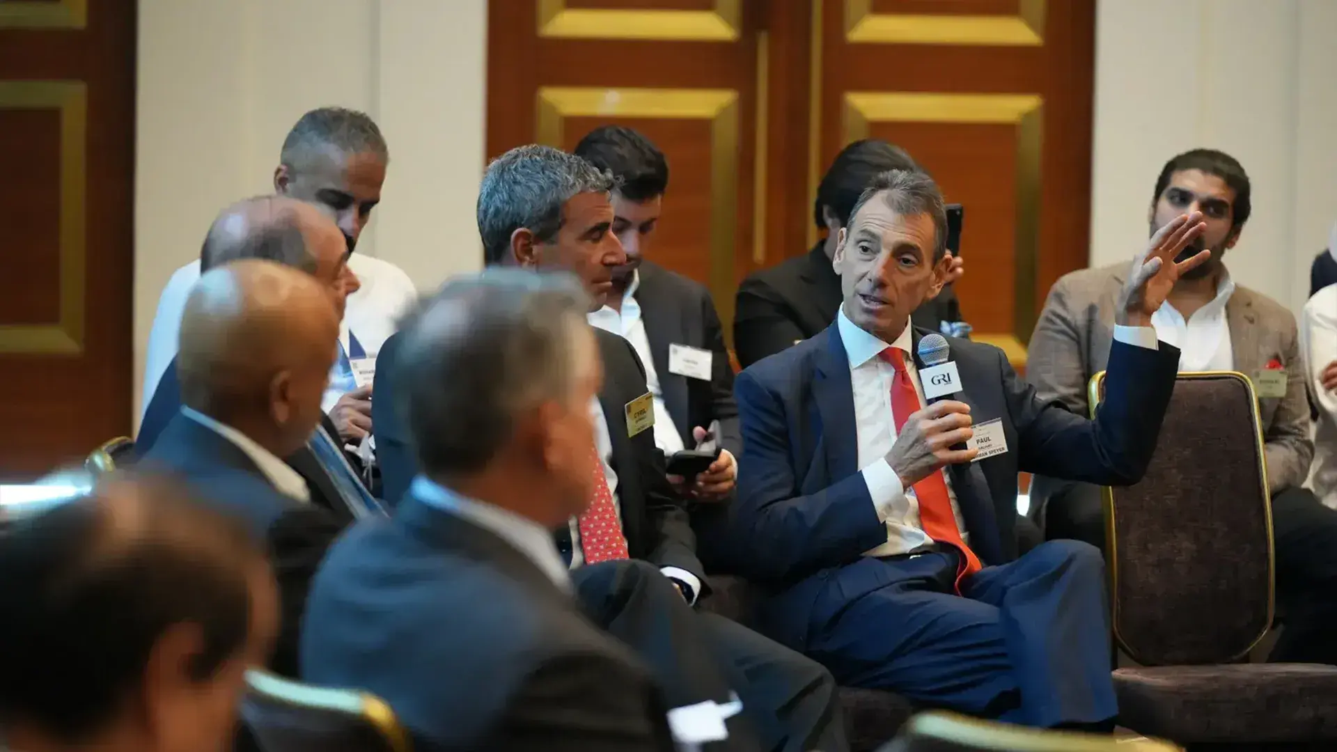A man in a navy suit and orange tie speaks animatedly into a microphone at a table during a global real estate summit, surrounded by other business professionals.