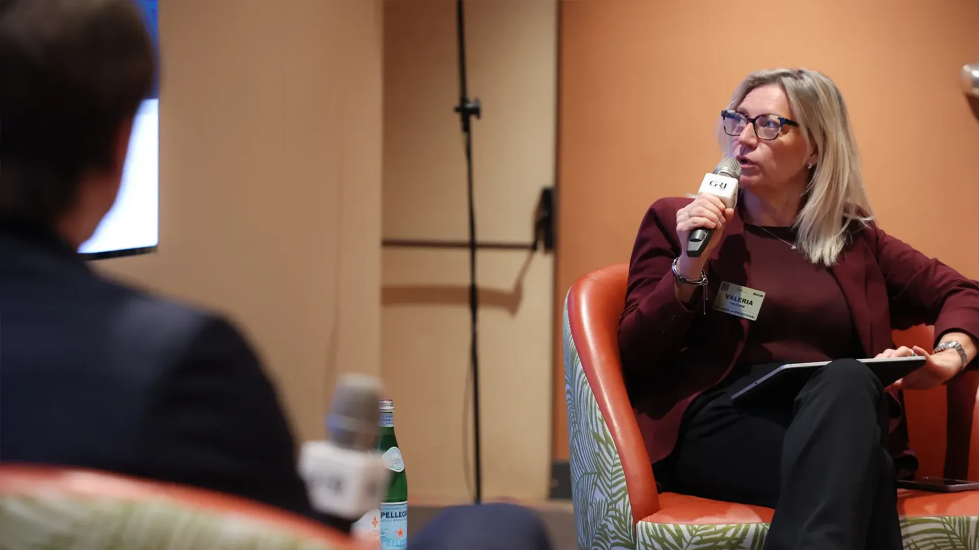 A female speaker, "Valeria," holding a microphone and addressing attendees while sitting in a large orange chair during a panel focused on hospitality real estate investment.