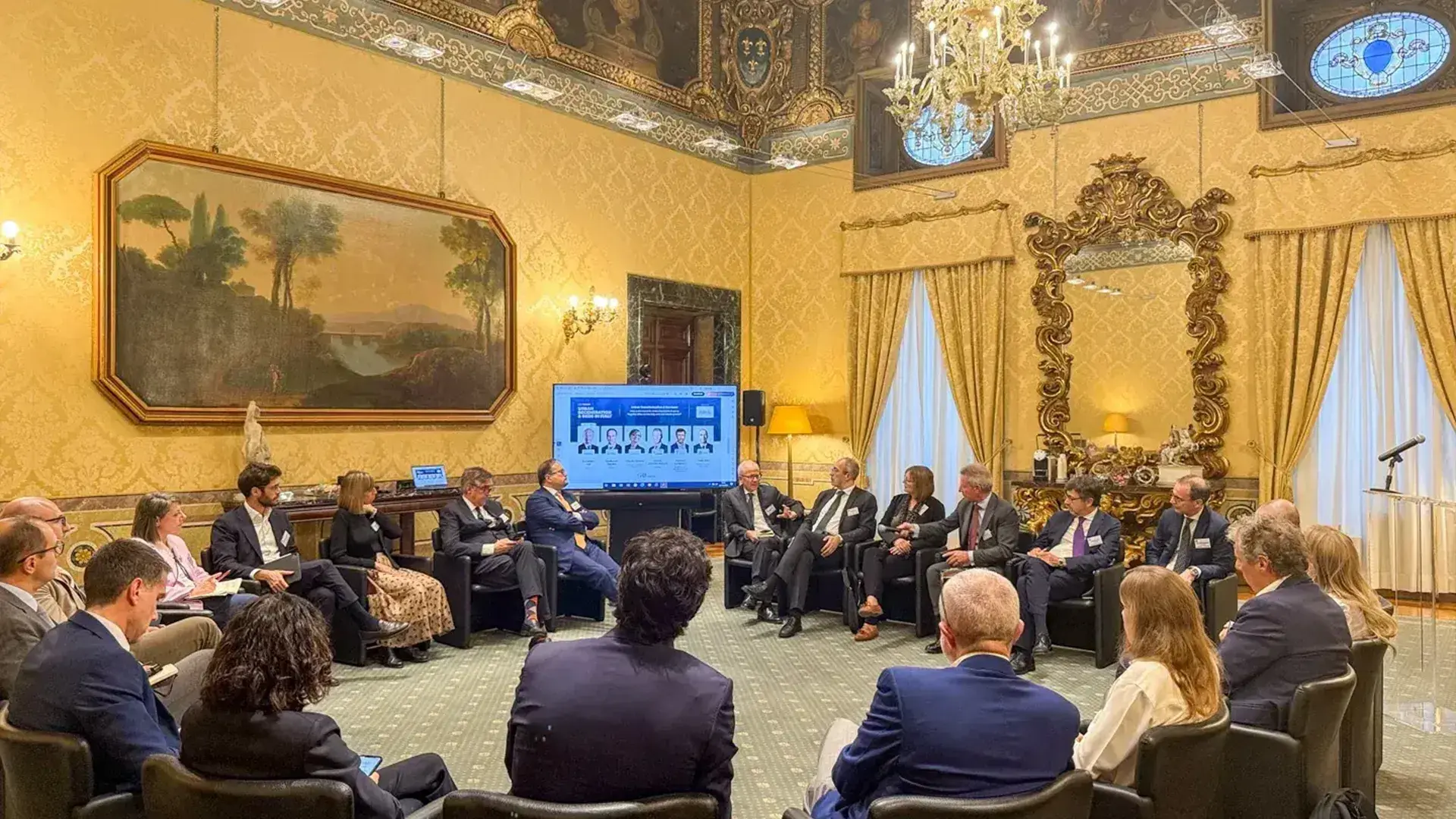 A group of real estate professionals is seated in a semi-circle in a luxuriously decorated, ornate room for a discussion, with a large screen displaying speaker information visible behind the attendees, relevant to the topic of Urban Regeneration & Beds in Italy.
