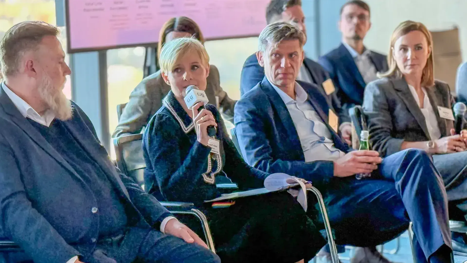 Panel discussion at a real estate conference, featuring a woman speaking into a microphone and surrounding attendees in Poland.