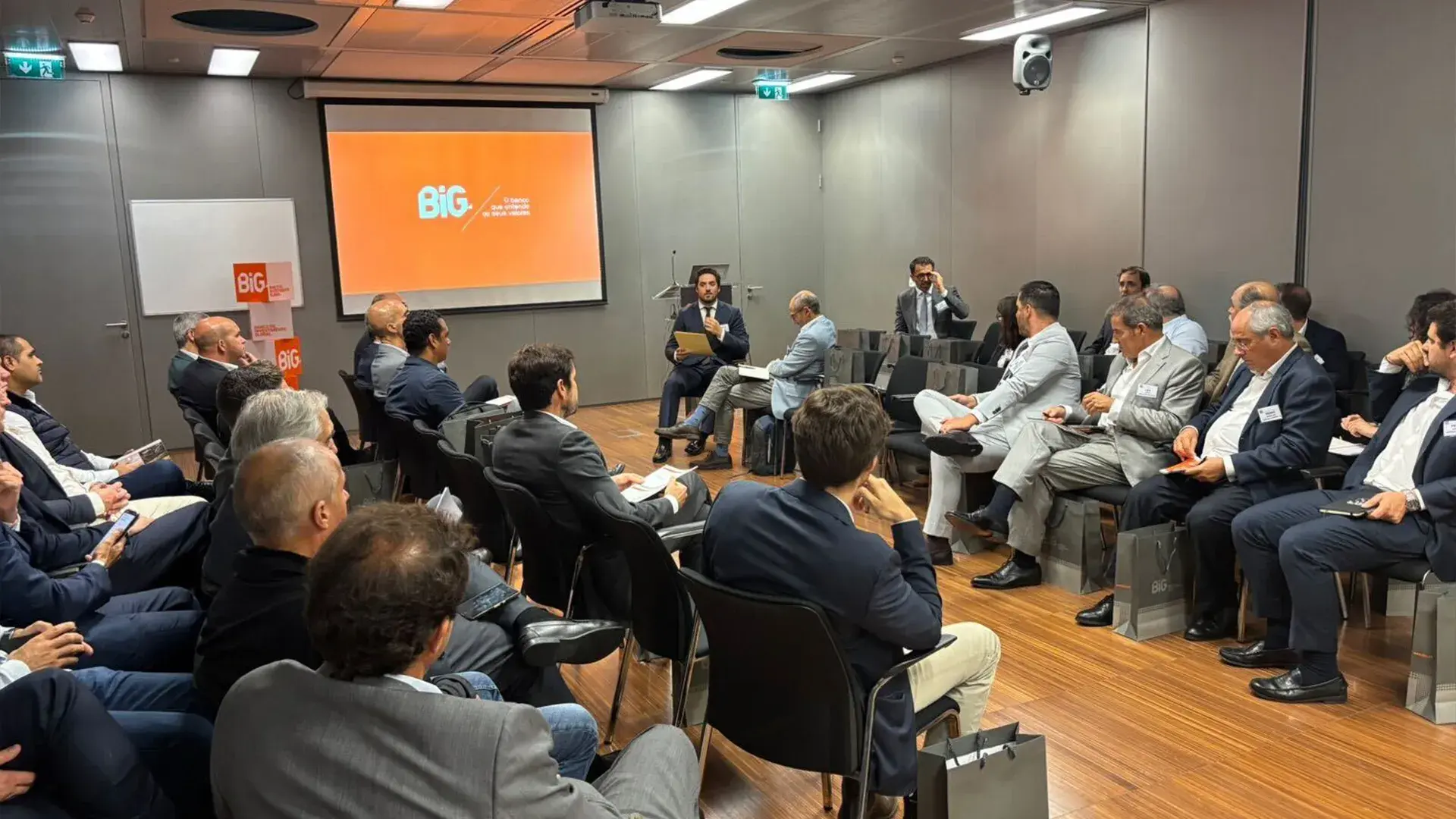 A group of business professionals attending a conference, discussing Portugal real estate and alternative finance, as suggested by the setting and overall context. A screen displays a logo featuring "BiG".