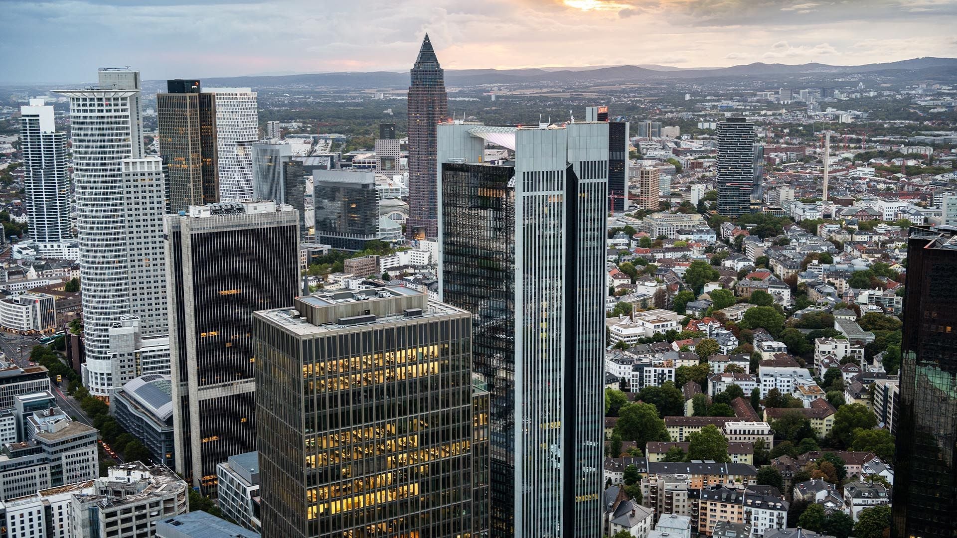 An aerial perspective of Frankfurt’s central business district at dusk. The skyline is dominated by high-rise office towers, including the Commerzbank Tower and Messeturm, interspersed with dense clusters of commercial and residential buildings characteristic of Germany’s financial capital.