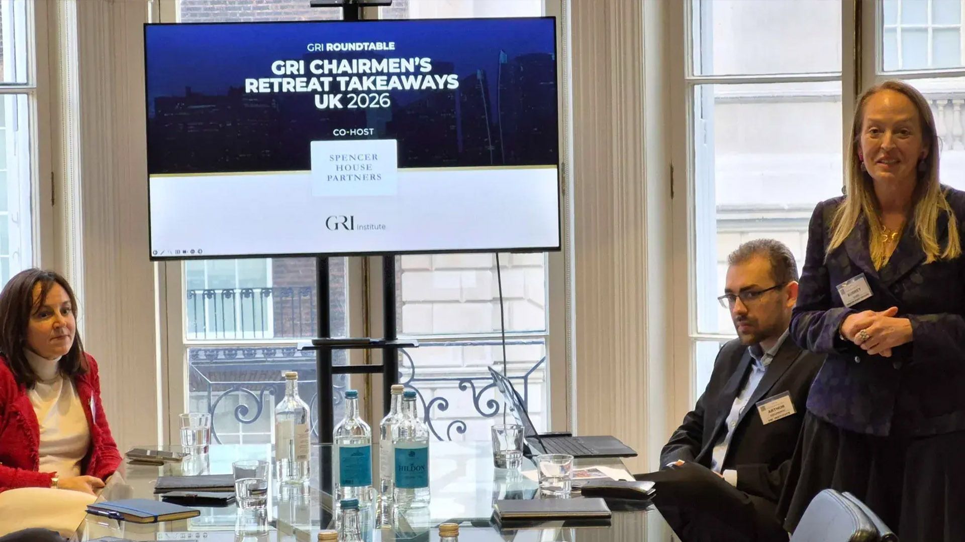 A close-up of a real estate industry presentation titled "GRI Chairmen's Retreat Takeaways UK 2026." A female speaker leads the discussion alongside a screen co-hosted by Spencer House Partners, while attendees listen at a glass boardroom table.