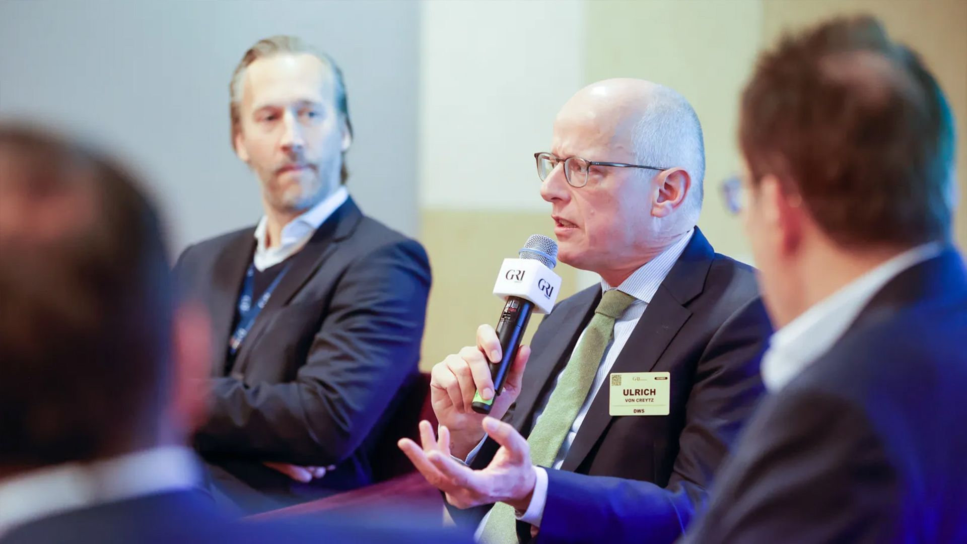 A bald man wearing glasses, a suit, and a light green tie, identified by his name badge as "Ulrich," is speaking into a microphone during a seated panel or conference. He is gesturing with his hands and is likely discussing topics related to the real estate market or investments in Germany.