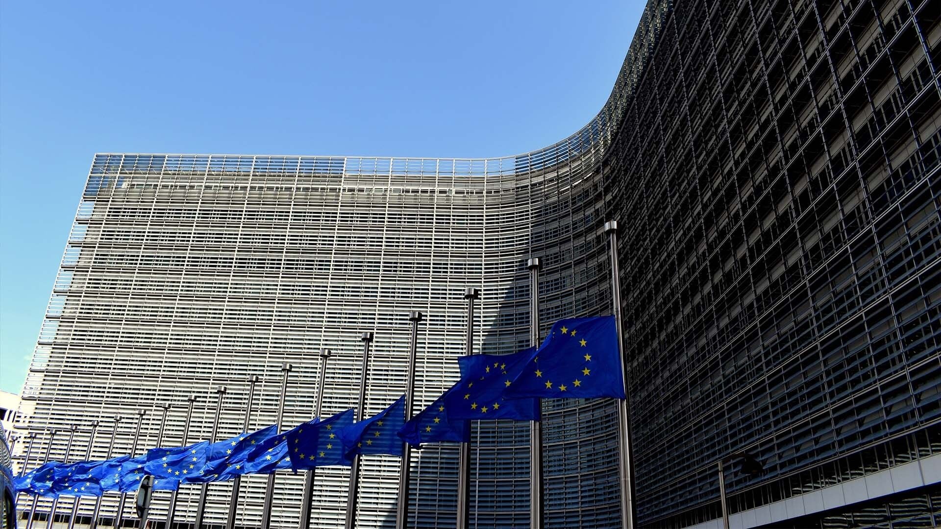 A row of European Union flags waving in front of the modern, glass-facaded Berlaymont building, representing the central hub for regulation and policy-making affecting the real estate sector across Europe.