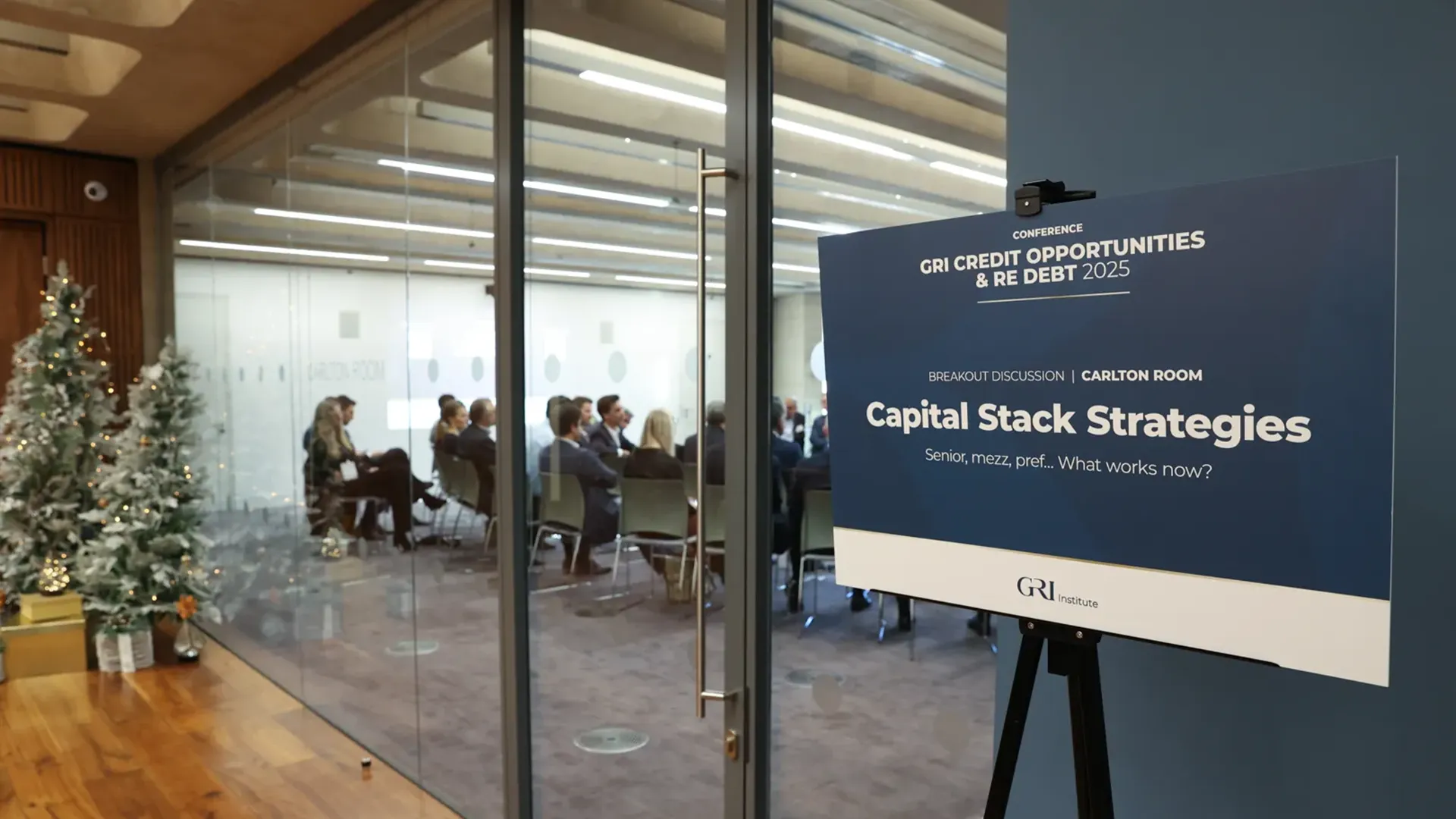 A conference sign for the GRI Credit Opportunities & RE Debt 2025 event is prominently displayed, detailing a Breakout Discussion | Carlton Room titled "Capital Stack Strategies." The subtitle reads: "Senior, mezz, pref... What works now?" The sign is on an easel next to a glass-walled room where a group of people are seated, facing forward, attending the discussion, with a decorated Christmas tree visible to the left. The scene captures a professional discussion related to real estate finance and debt.