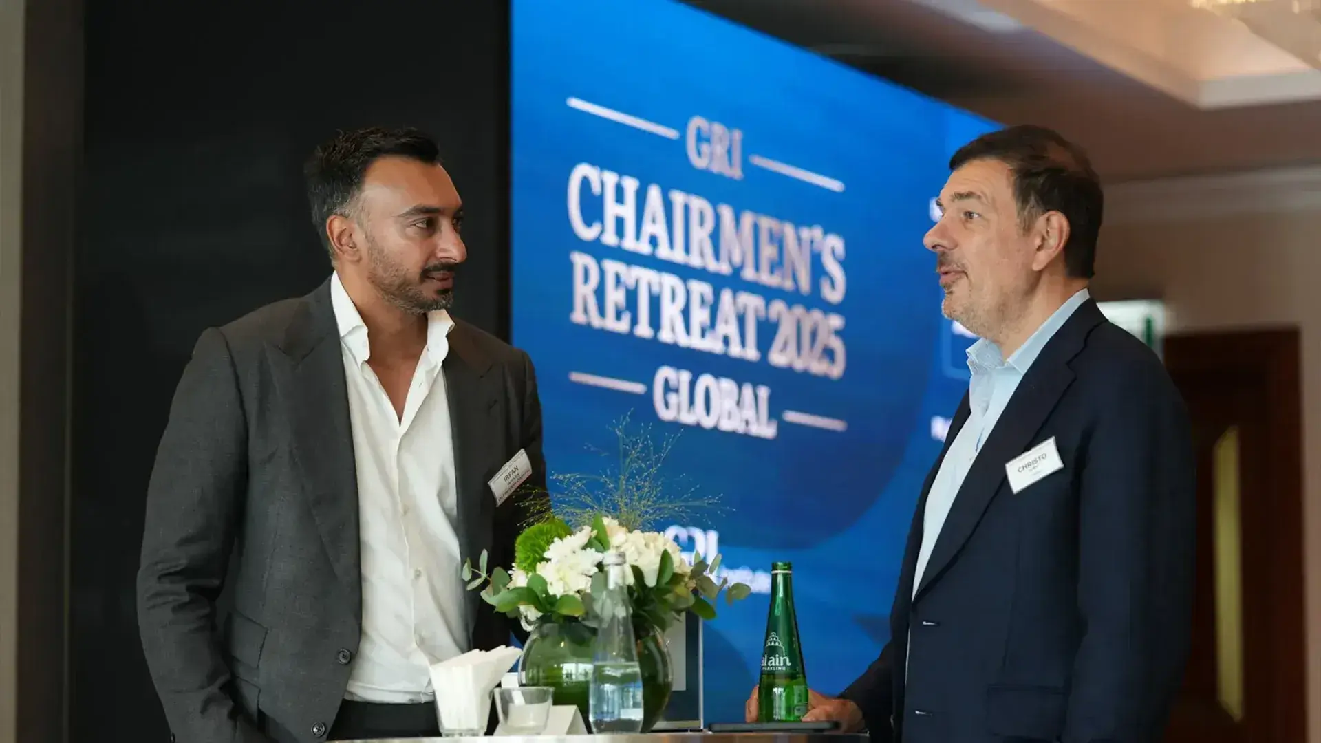 Two global real estate leaders are engaged in a side conversation during the GRI Global Chairmen's Retreat 2025, with a display screen visible behind them.