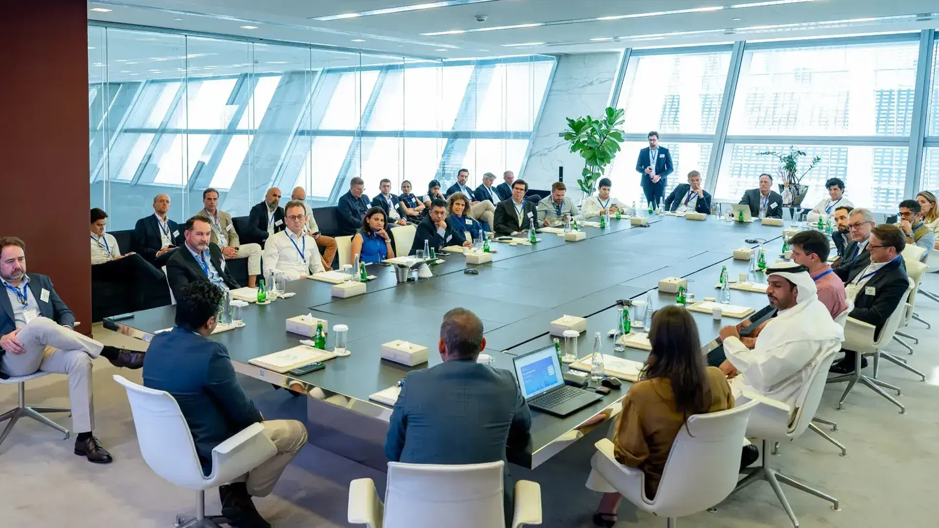 A large group of executives and business professionals attending a meeting around a long black table in a modern, glass-walled conference room at ADGM (Abu Dhabi Global Market). The group is likely discussing topics such as finance, investment, or real estate.