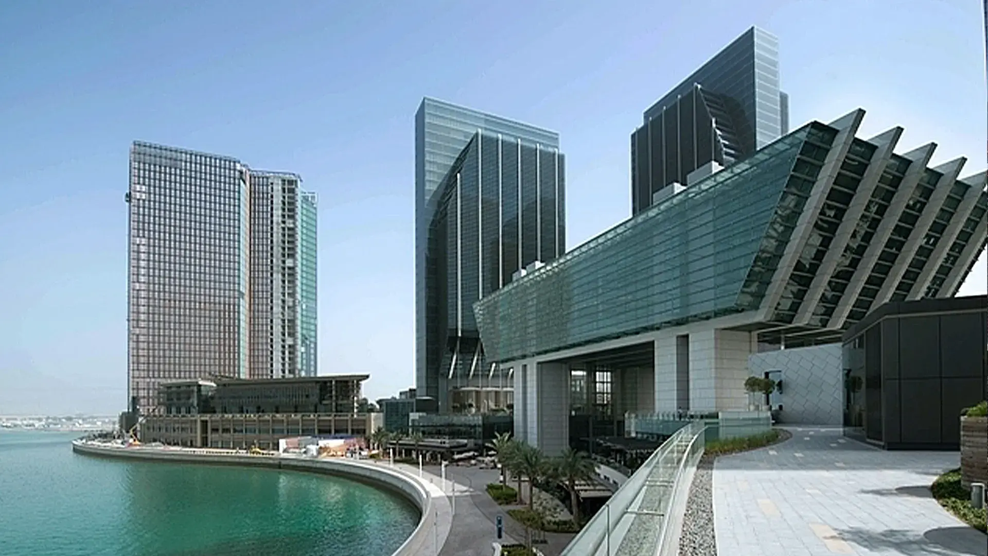 A wide shot of the Abu Dhabi Global Market (ADGM) complex, featuring modern, glass high-rise towers and a distinctive glass-enclosed, elevated structure, all set along a curved waterfront.