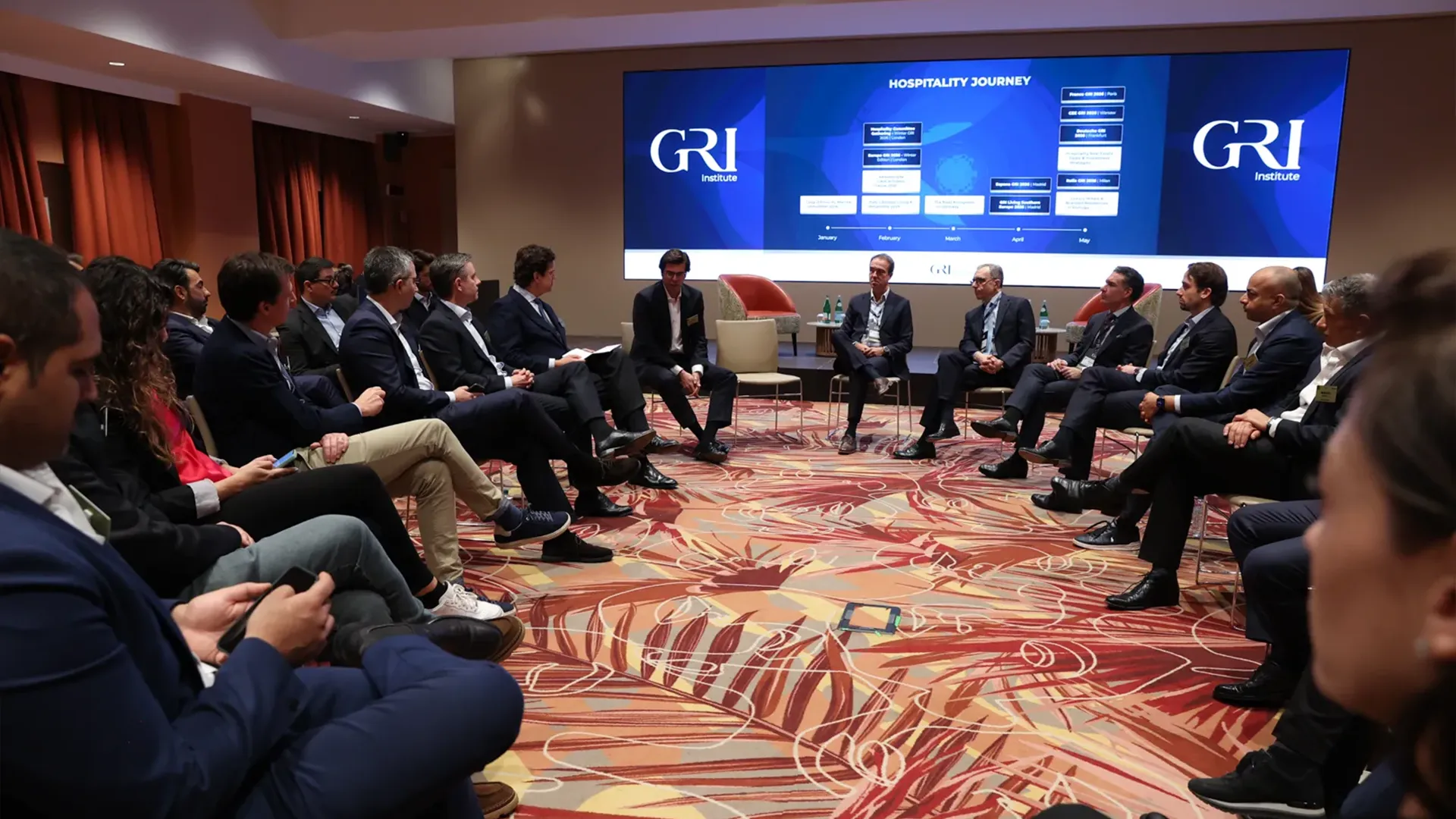 A group of executives seated in a semi-circle around a small panel of speakers, looking toward a large screen displaying a "Hospitality Journey" timeline at the GRI Institute event on real estate development.