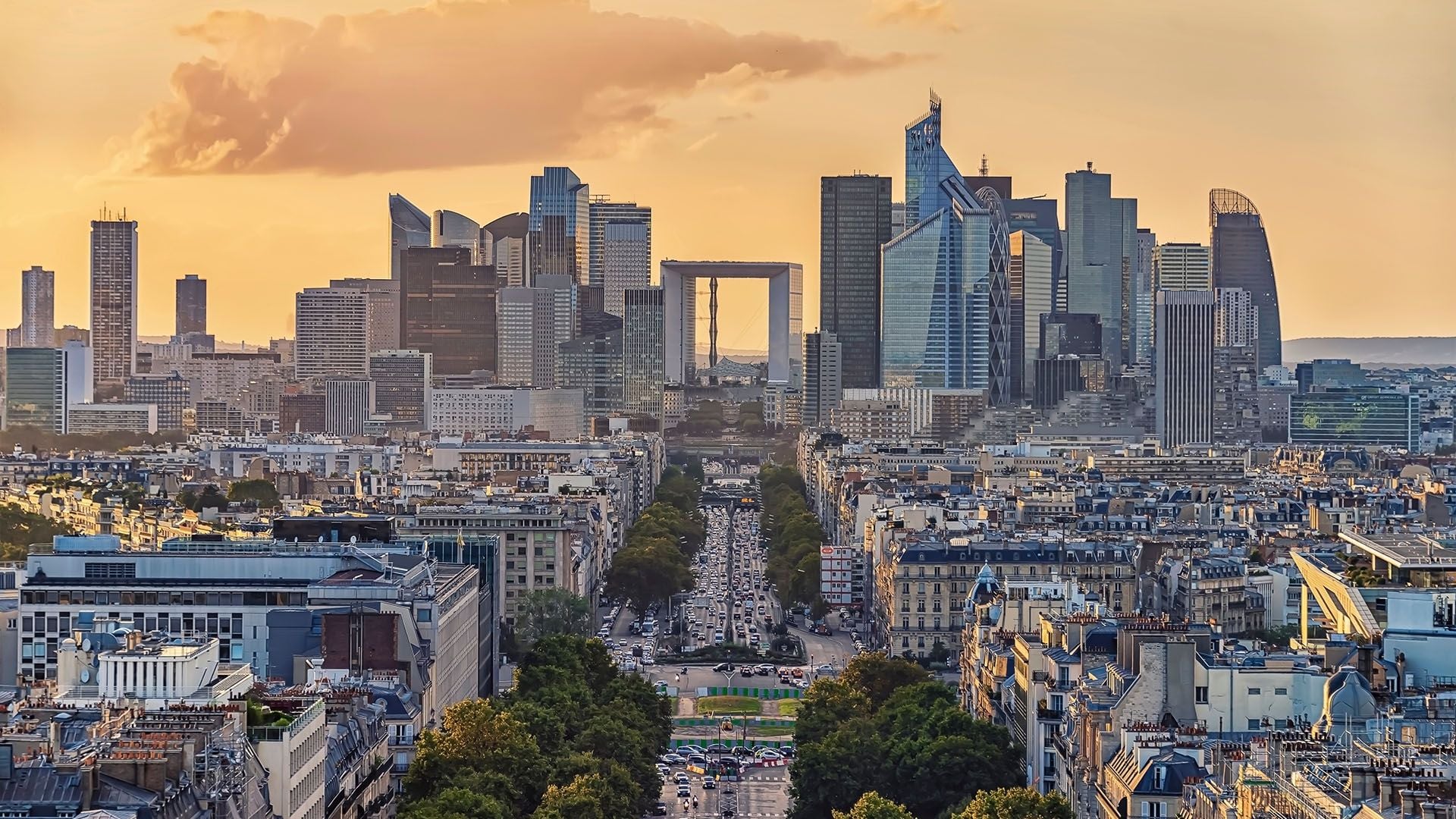 A sunset view looking down the Avenue de la Grande Armée toward the La Défense business district. The Grande Arche de la Défense is framed by a symmetrical alignment of modern office towers and classic Haussmann-style Parisian apartment buildings in the foreground.