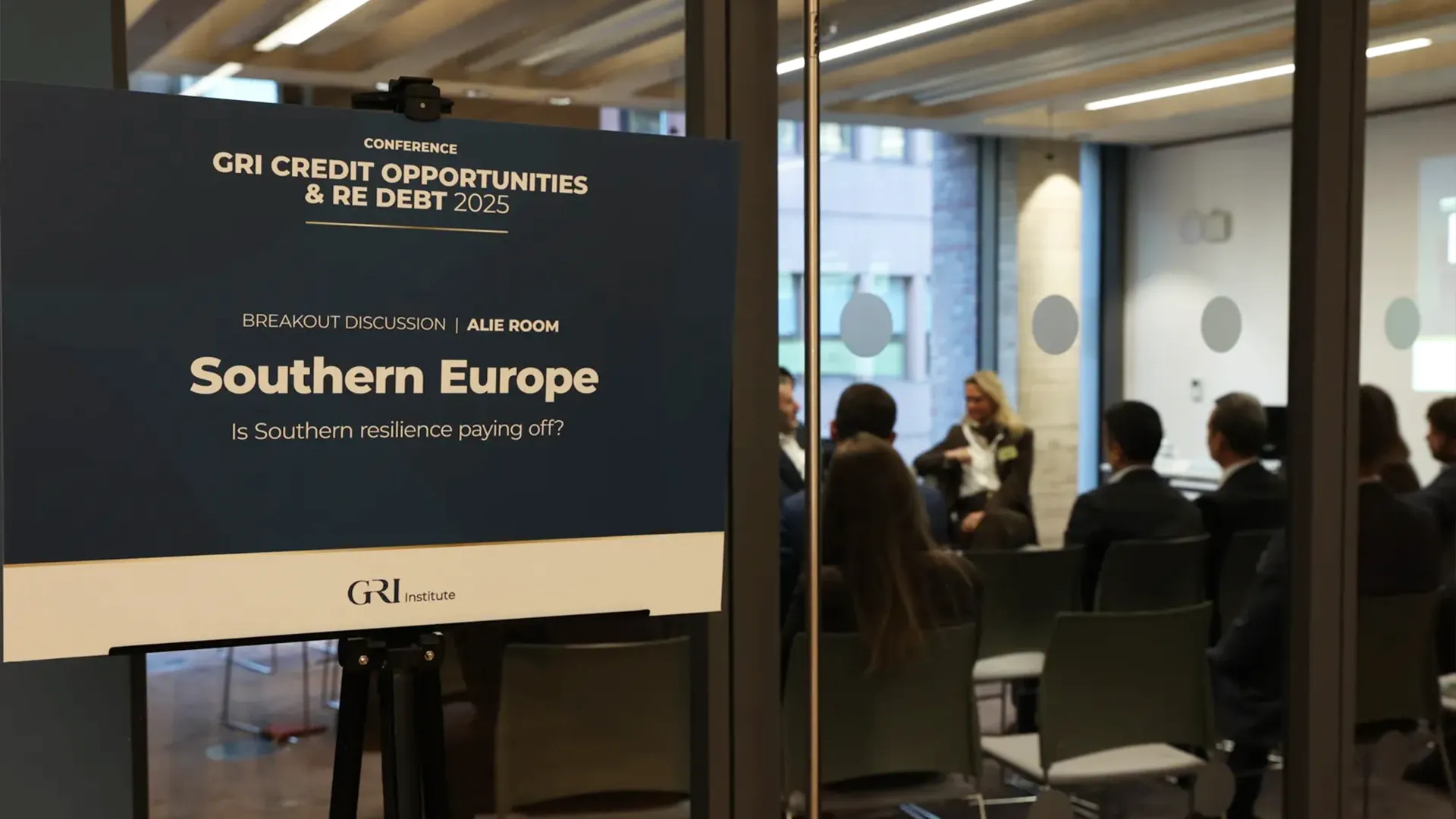 A large sign for the GRI Credit Opportunities & RE Debt 2025 conference is positioned in front of a glass-walled room where a breakout discussion is taking place. The logo for GRI Institute is at the bottom of the sign. Through the glass, several people in business attire are visible, seated and engaged in the discussion, with a woman speaking in the center. The image captures a moment from a financial conference focused on real estate debt and opportunities.