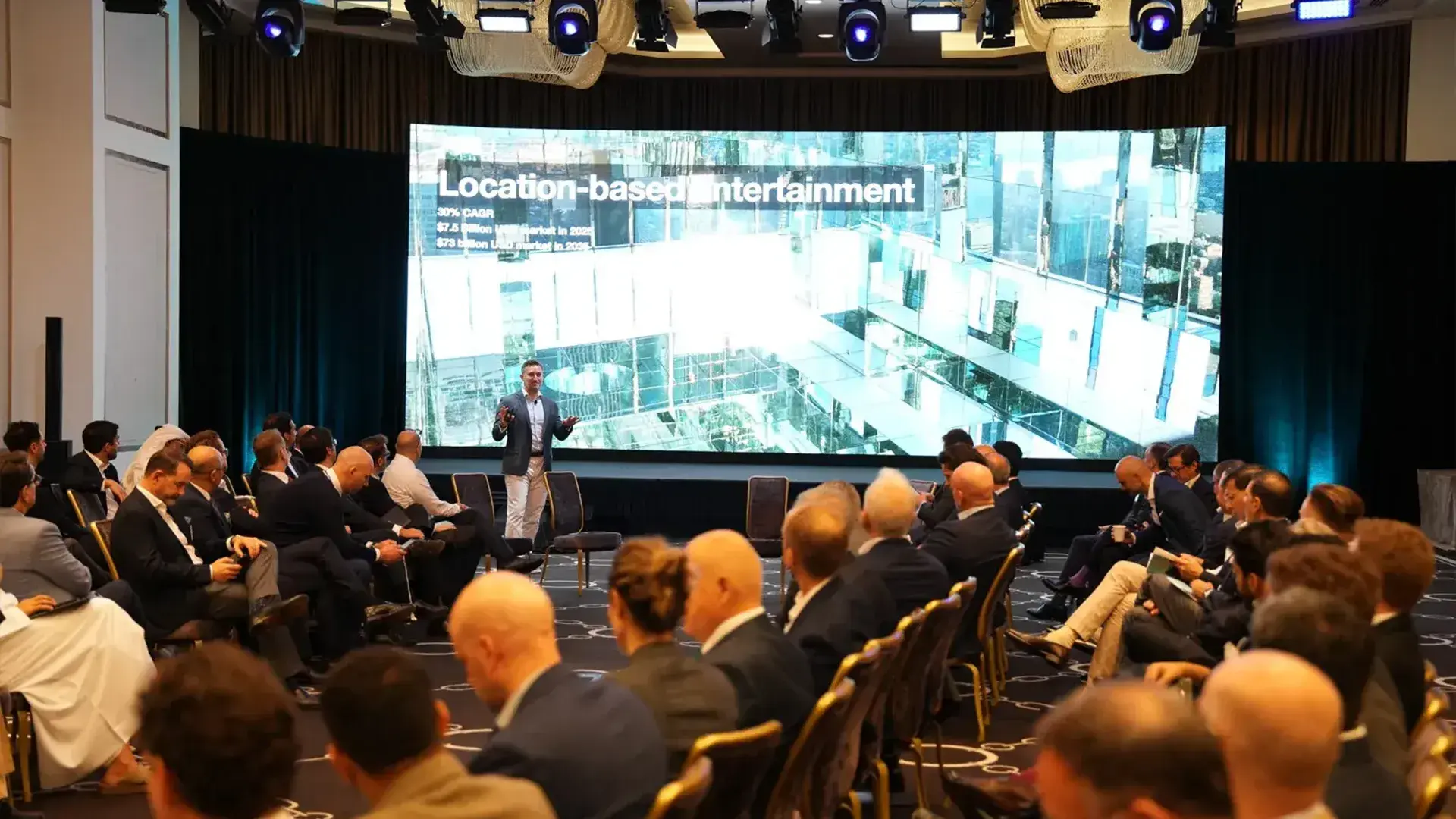 A presenter speaks on stage about "Location-based Entertainment" to an audience of global real estate leaders at a conference. The large screen displays market data and a modern building image.