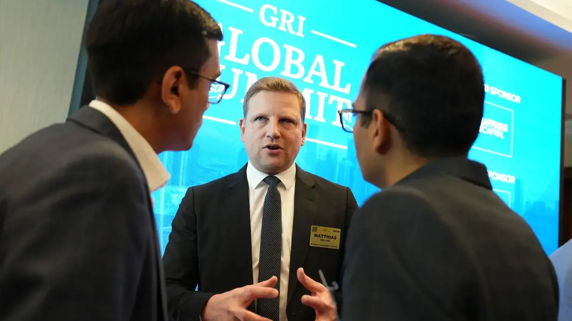 A professional man in a suit, wearing a name tag that reads "MATTHIAS," speaks intently to two other men on either side of him at a global summit. A large blue screen in the background features the text "GRI GLOBAL SUMMIT" and mentions a sponsor, relating to the real estate industry.