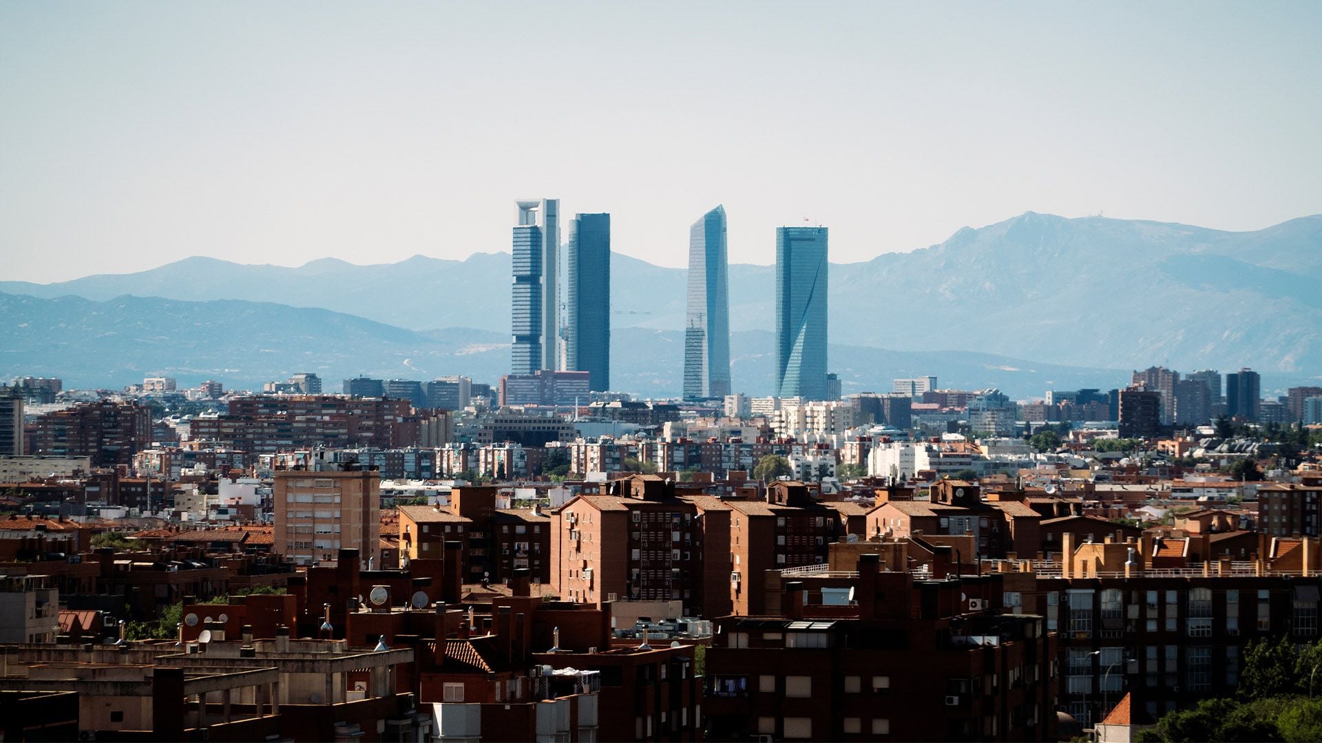 A distant view of the Cuatro Torres Business Area, Madrid’s primary cluster of modern skyscrapers, rising above a vast expanse of traditional brick residential blocks. The snow-capped Guadarrama Mountains provide a sharp natural backdrop to the city's northern business hub.