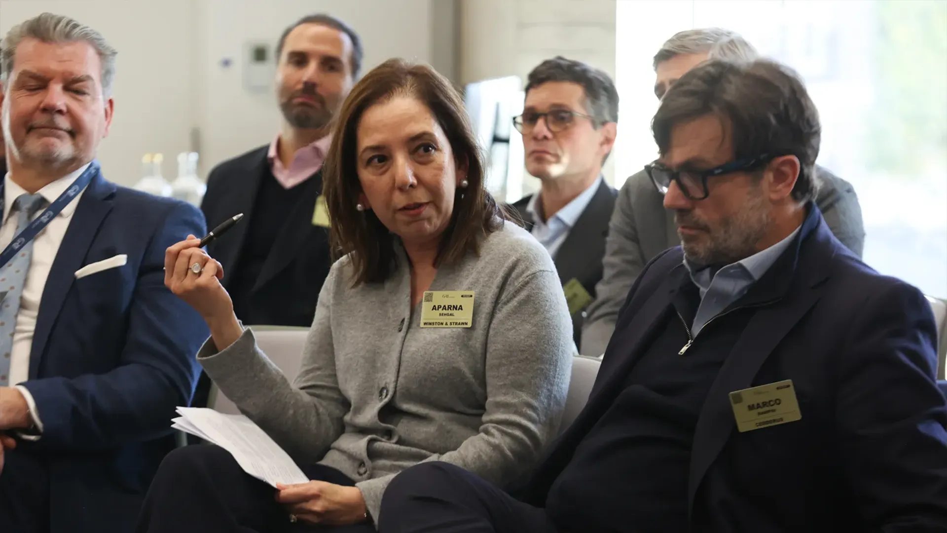 A woman in a grey cardigan, identified by a badge as Aparna, from 'Winston & Strawn', is speaking and holding a pen, while seated among several other professionals in business attire at a conference or meeting. A man in the foreground to her right, wearing glasses and a dark blue jacket, is identified as Marco. The setting appears to be an indoor event, likely related to real estate given the context of professional discussions.