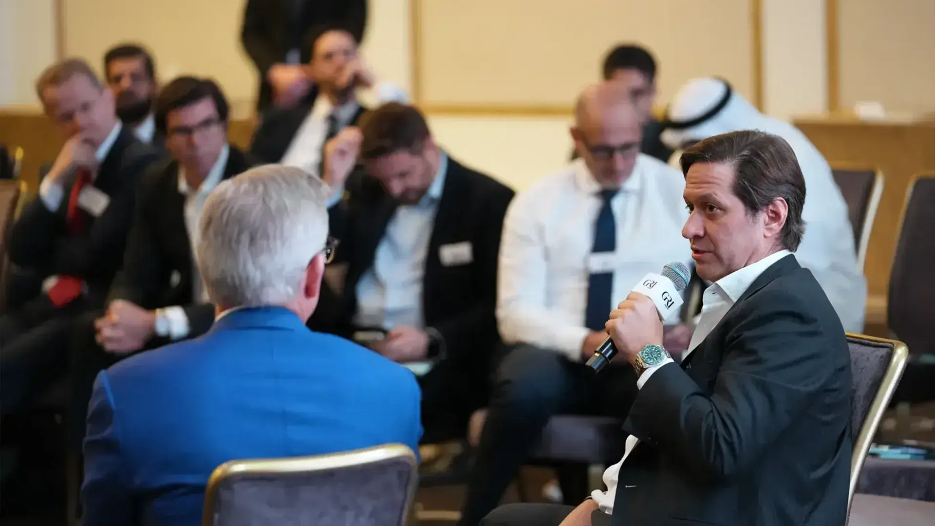 A focused session featuring global real estate leaders, with one man in the foreground speaking into a microphone while others in the audience listen attentively.