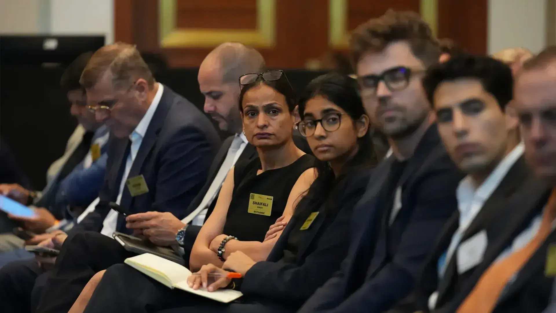 Attendees, including a woman with a name tag visible reading 'SHAIFALI', are seated and attentively listening at a session during a global real estate summit. Several people are dressed in business attire and many are looking towards the front of the room.