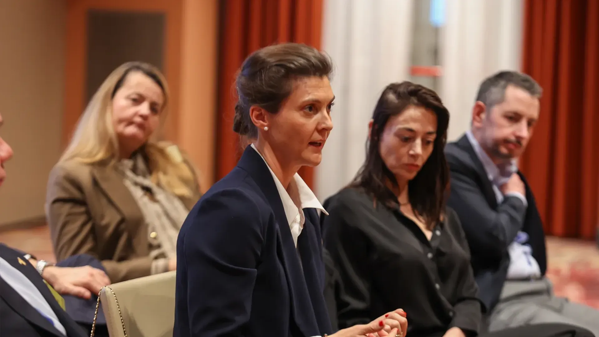 A female executive in a navy blazer and white shirt speaking to a group of peers during a discussion about real estate asset management within the hospitality industry.