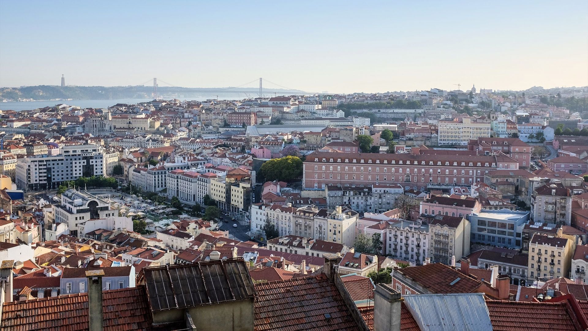 A high-angle, panoramic view of Lisbon’s dense urban fabric, featuring traditional red-tiled roofs in the foreground. The city slopes down toward the Tagus River, where the 25 de Abril Bridge is visible on the horizon. The architecture is a mix of historic Portuguese buildings and mid-century apartment blocks.