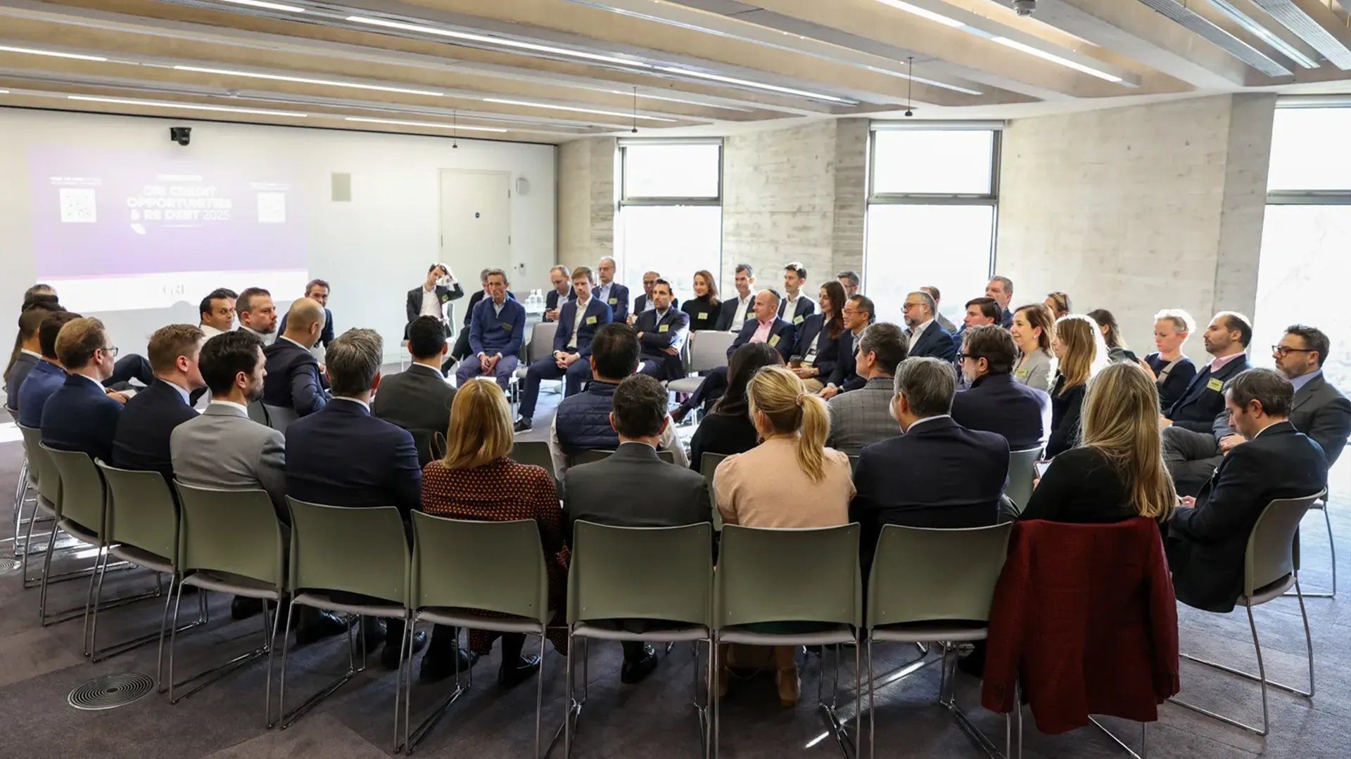 An audience of men and women, professionally dressed, are seated in a curved arrangement facing a panel of speakers and a projection screen in a modern, well-lit conference room. The screen displays the text "THE GREATEST OPPORTUNITIES IN RE DEBT 2025." This image depicts a professional conference or seminar on real estate finance or investment.