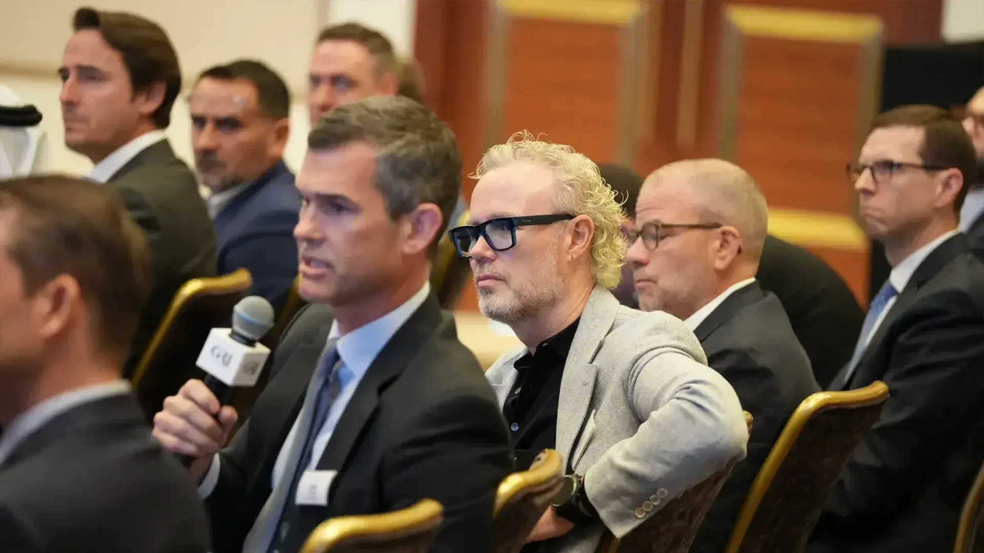  Alternative Text:  A line of attentive global real estate leaders sitting in chairs during a conference, with one man holding a microphone in the foreground as others listen.