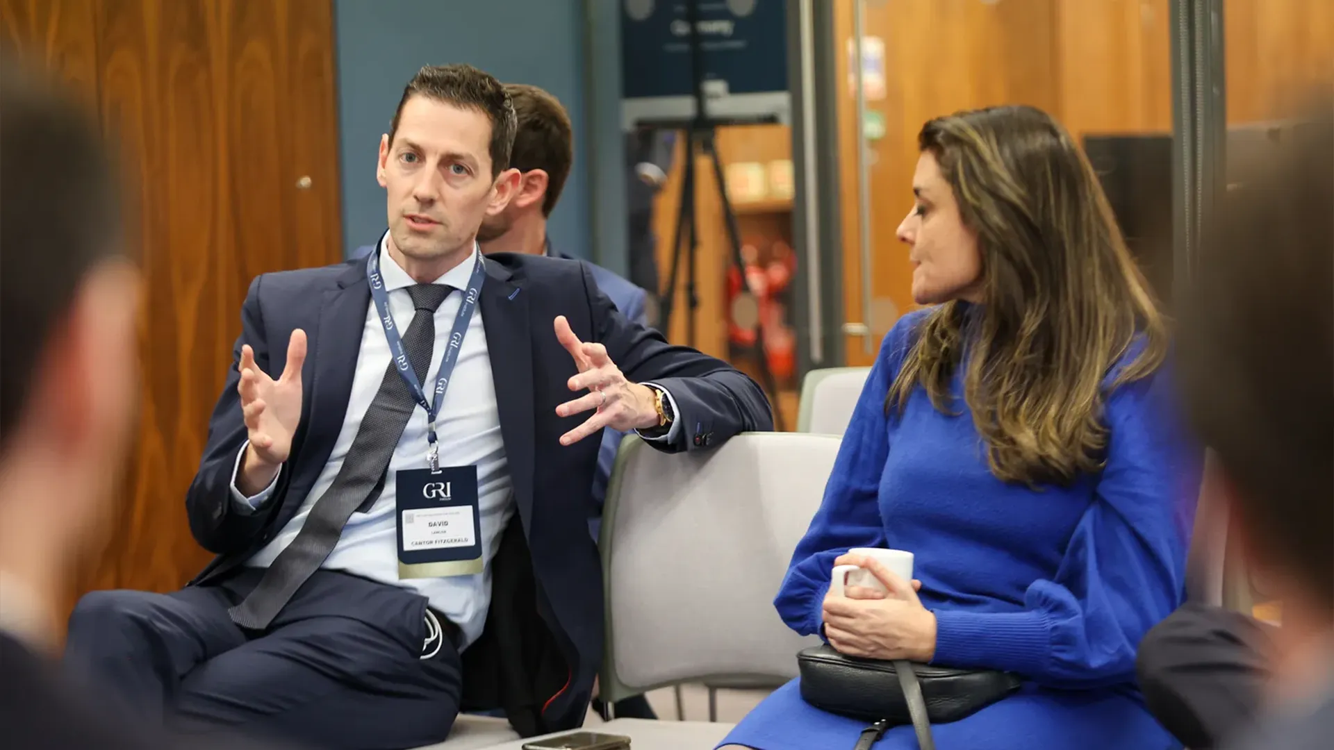 A man in a dark suit, wearing a real estate conference badge, speaks animatedly with his hands during a seated discussion with a woman in a royal blue dress.