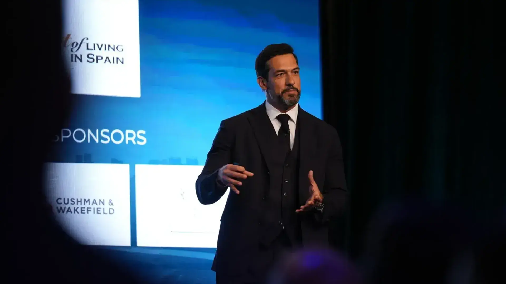 A speaker in a dark suit presenting on stage at a global real estate summit, with sponsor logos visible in the background.