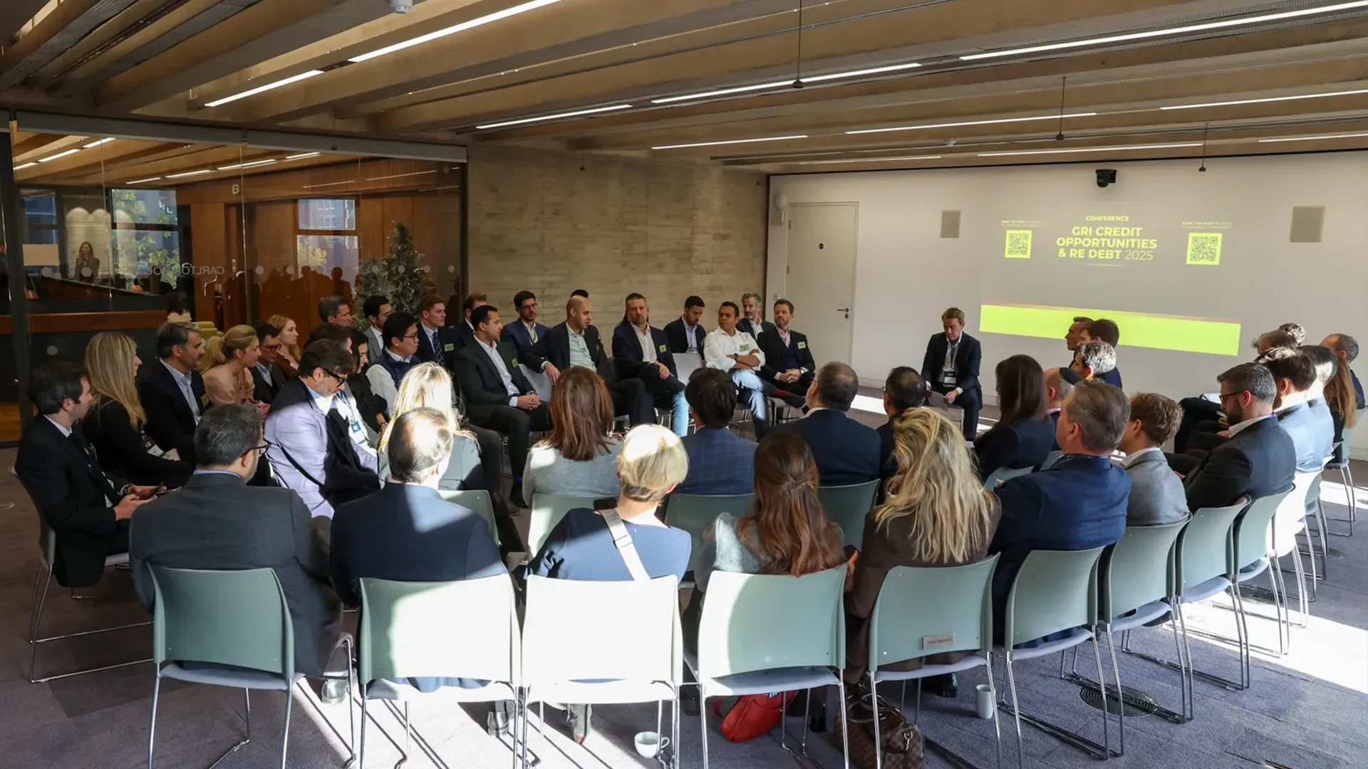 A group of approximately thirty people are seated in a semi-circle, facing a panel of seven men sitting on high stools at the front of a modern, brightly lit conference room. The panel members are dressed in business attire, some holding microphones, engaging in a discussion. On a large screen behind the panel, the text "CONFERENCE" is visible above the title "GRI CREDIT OPPORTUNITIES & RE DEBT 2025", with "RE DEBT" likely referring to real estate debt. Two QR codes are also displayed on the screen. The audience members, also in professional clothing, are attentive. The room has light-colored walls and a slatted wood ceiling.