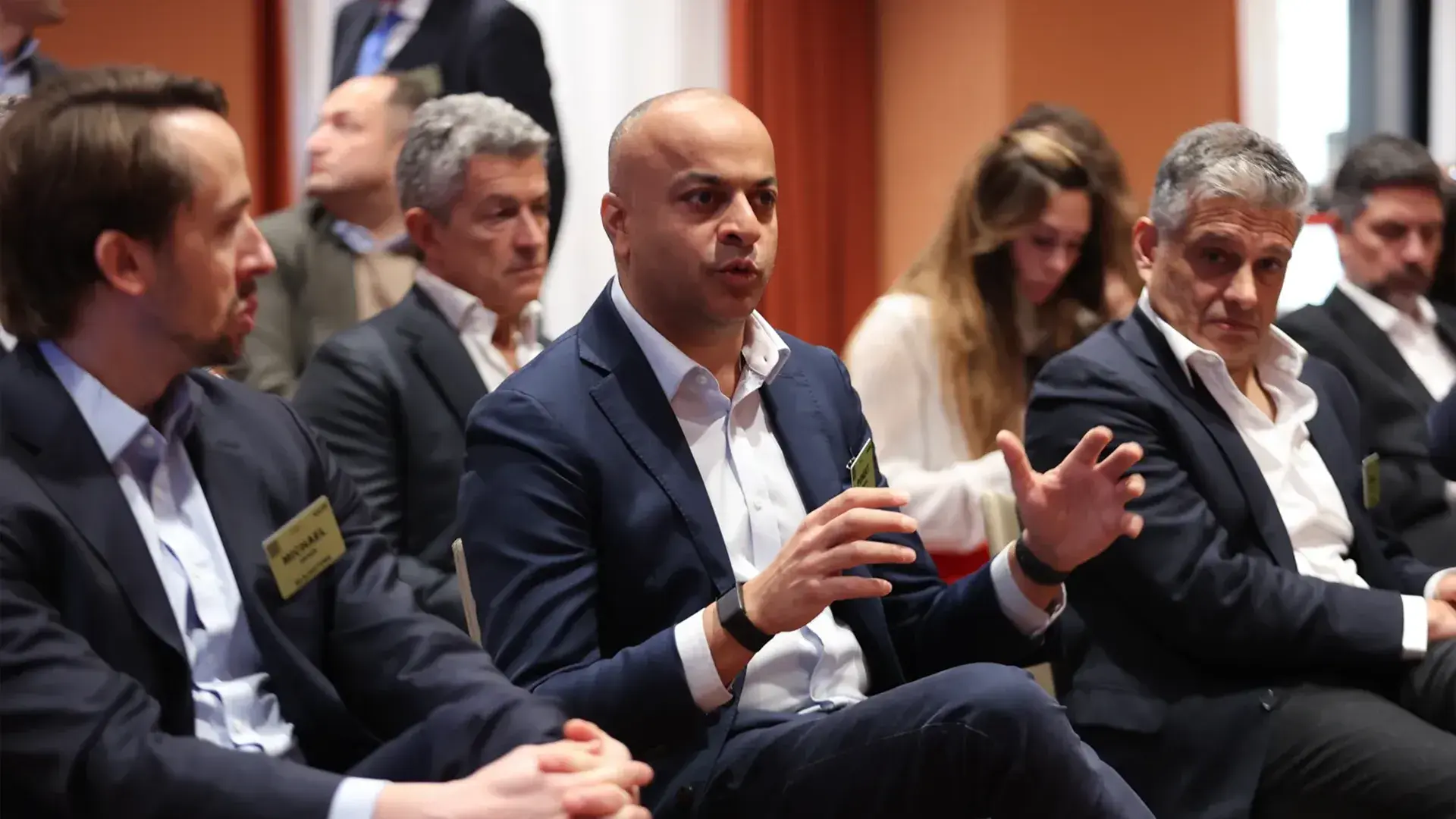 A business professional in a dark suit speaking animatedly with his hands during a focused group discussion on hospitality real estate during the GRI conference.