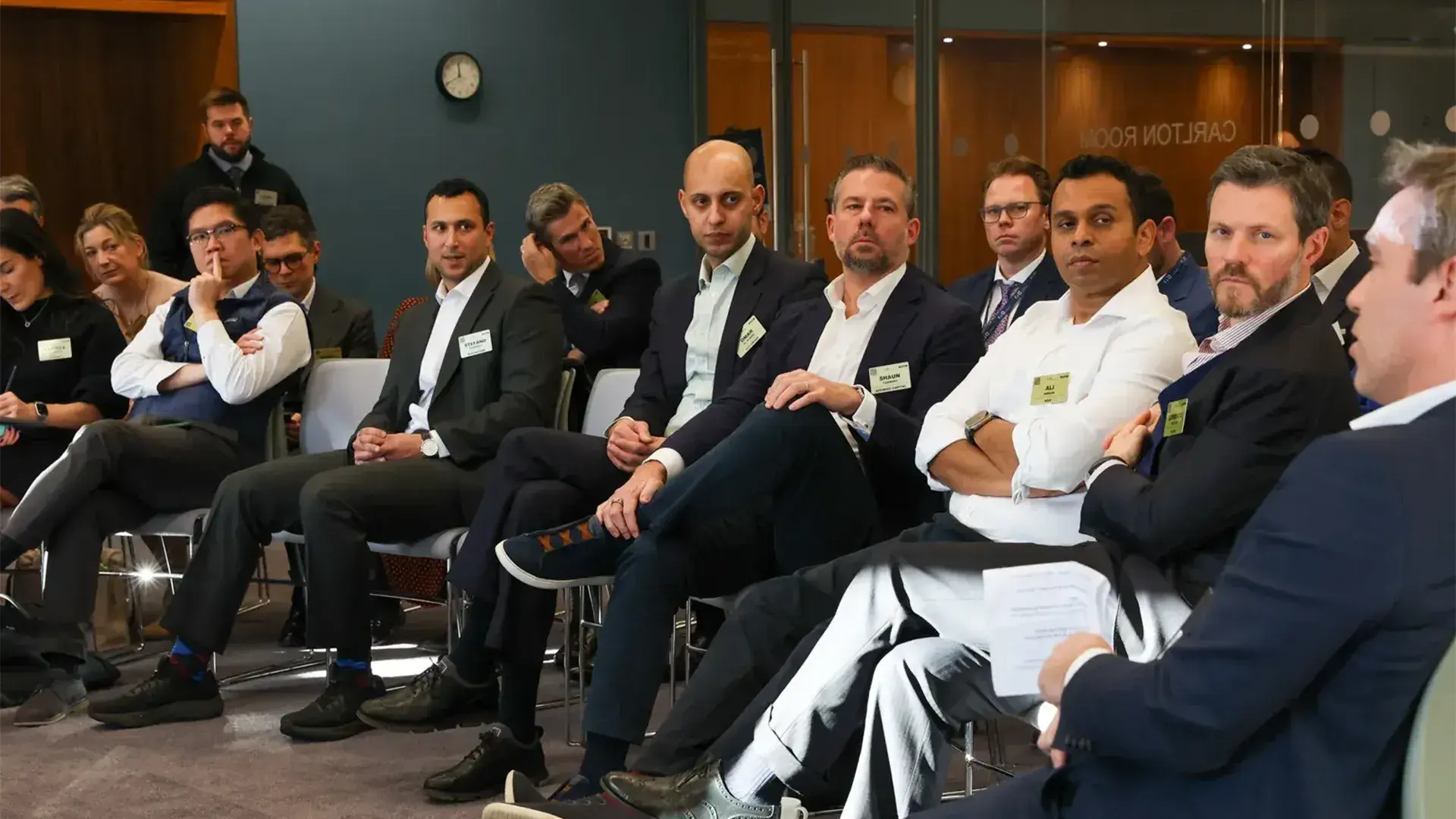 A group of professionals seated in a row at a real estate industry event hosted by the GRI Institute. The attendees, mostly dressed in business casual attire, are listening intently to a speaker (partially visible in the foreground) in a modern conference room with "Carlton Room" visible on a glass door in the background.